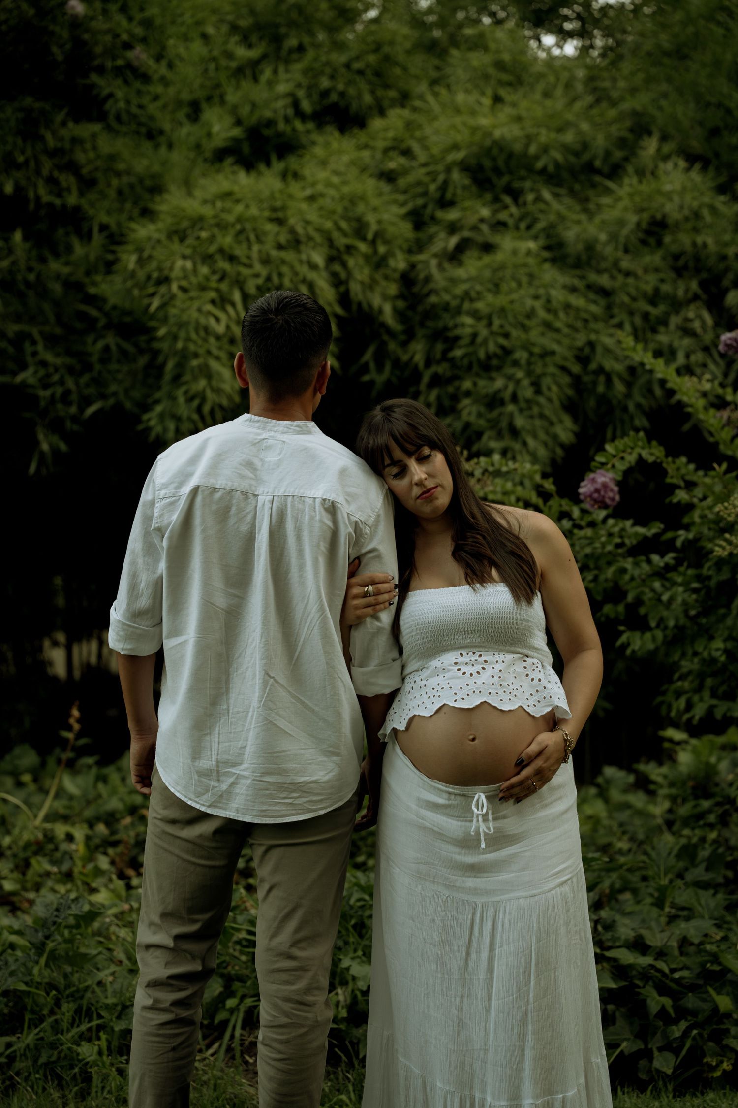 Pregnant woman in white dress standing beside partner in lush green garden, cradling her baby bump.