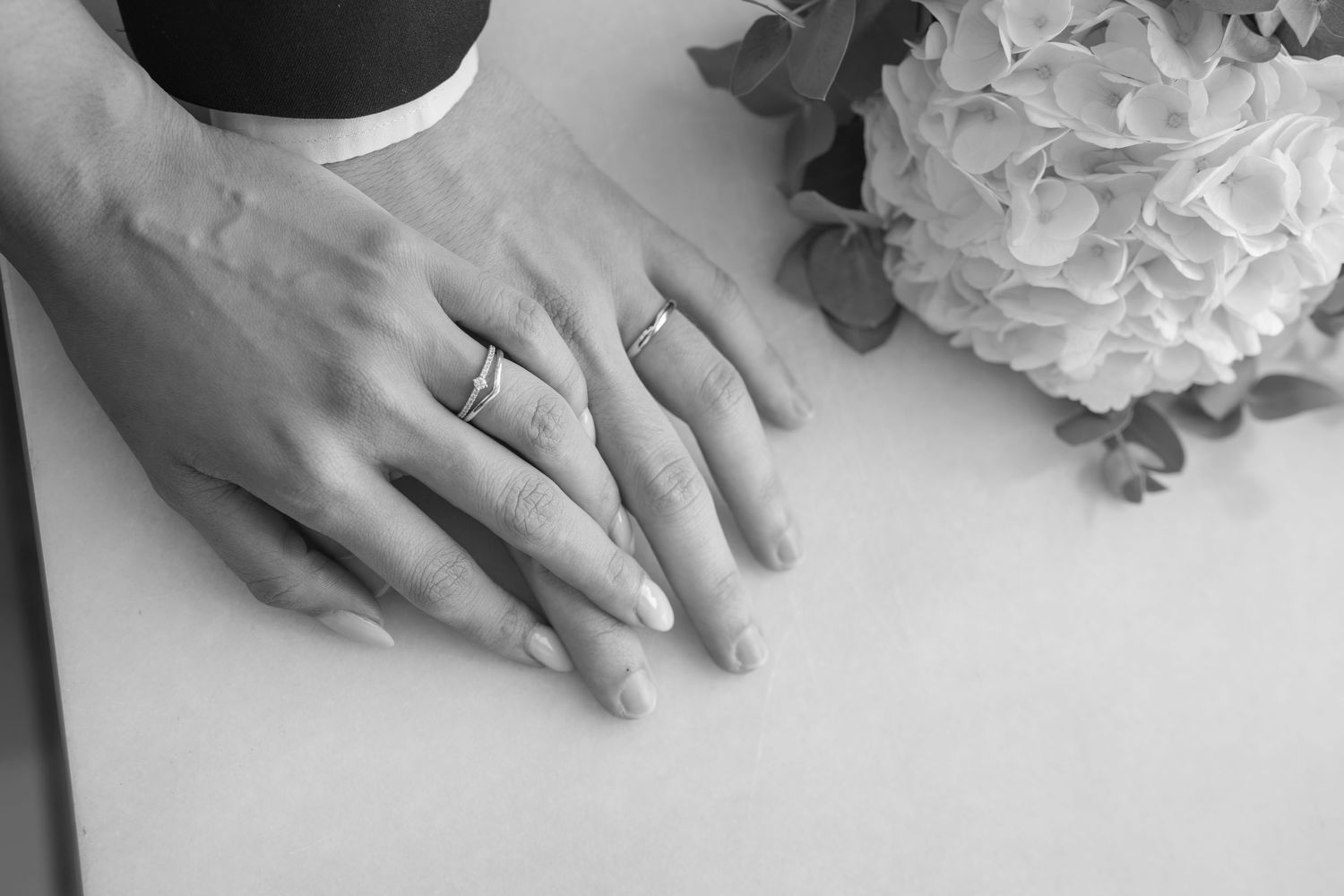 Black and white photo of two hands resting together wearing wedding rings beside a white hydrangea bouquet on a table.