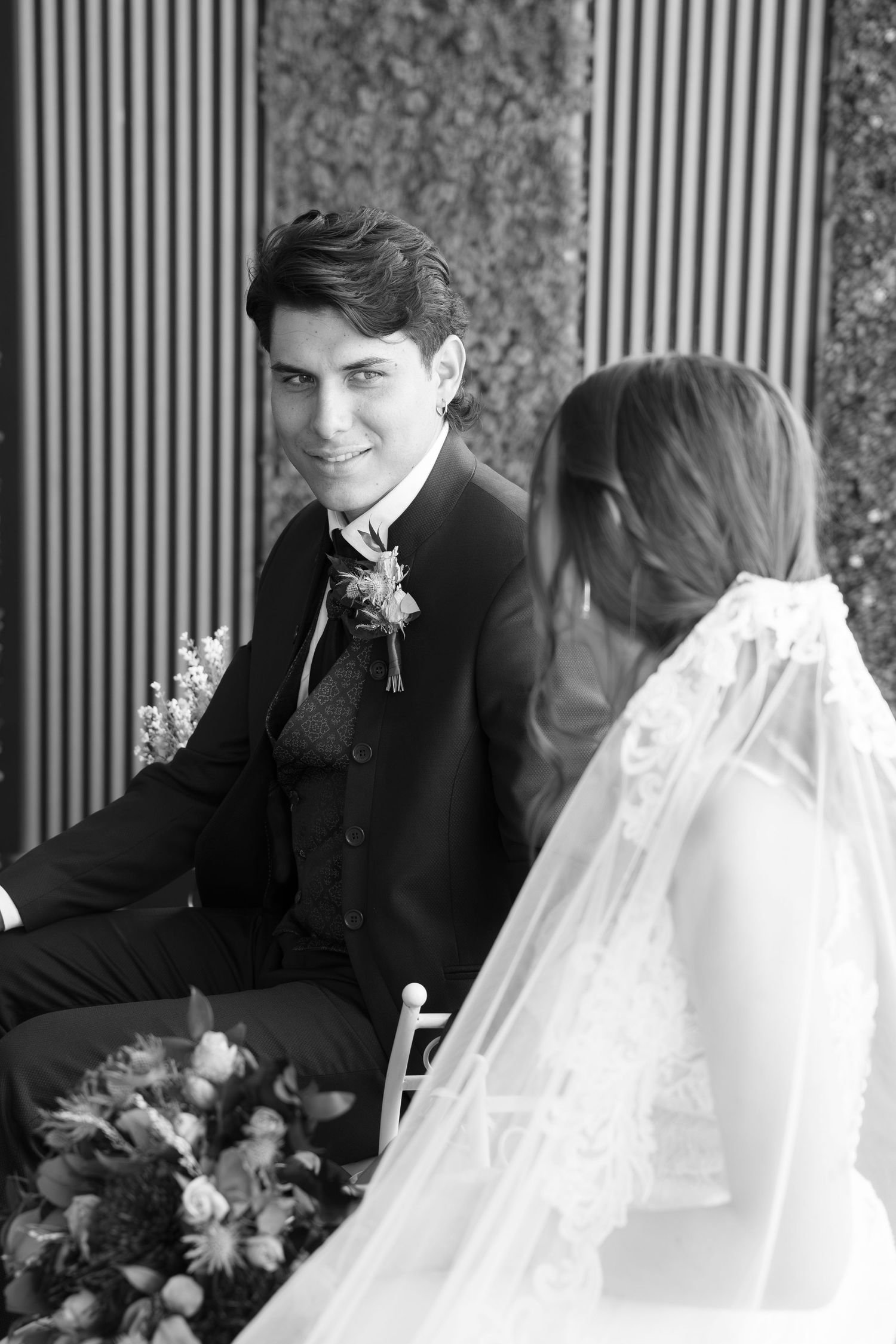 Black and white wedding photo of a smiling groom in a suit with boutonniere gazing at his veiled bride, with a floral bouquet nearby.