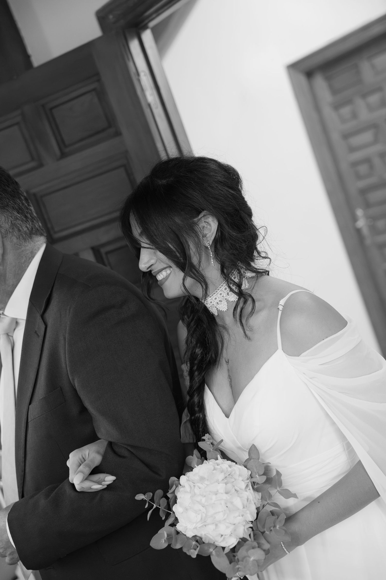 Smiling bride in a white off-shoulder gown with a braided updo holds a white bouquet while linking arms with a man in a dark suit.