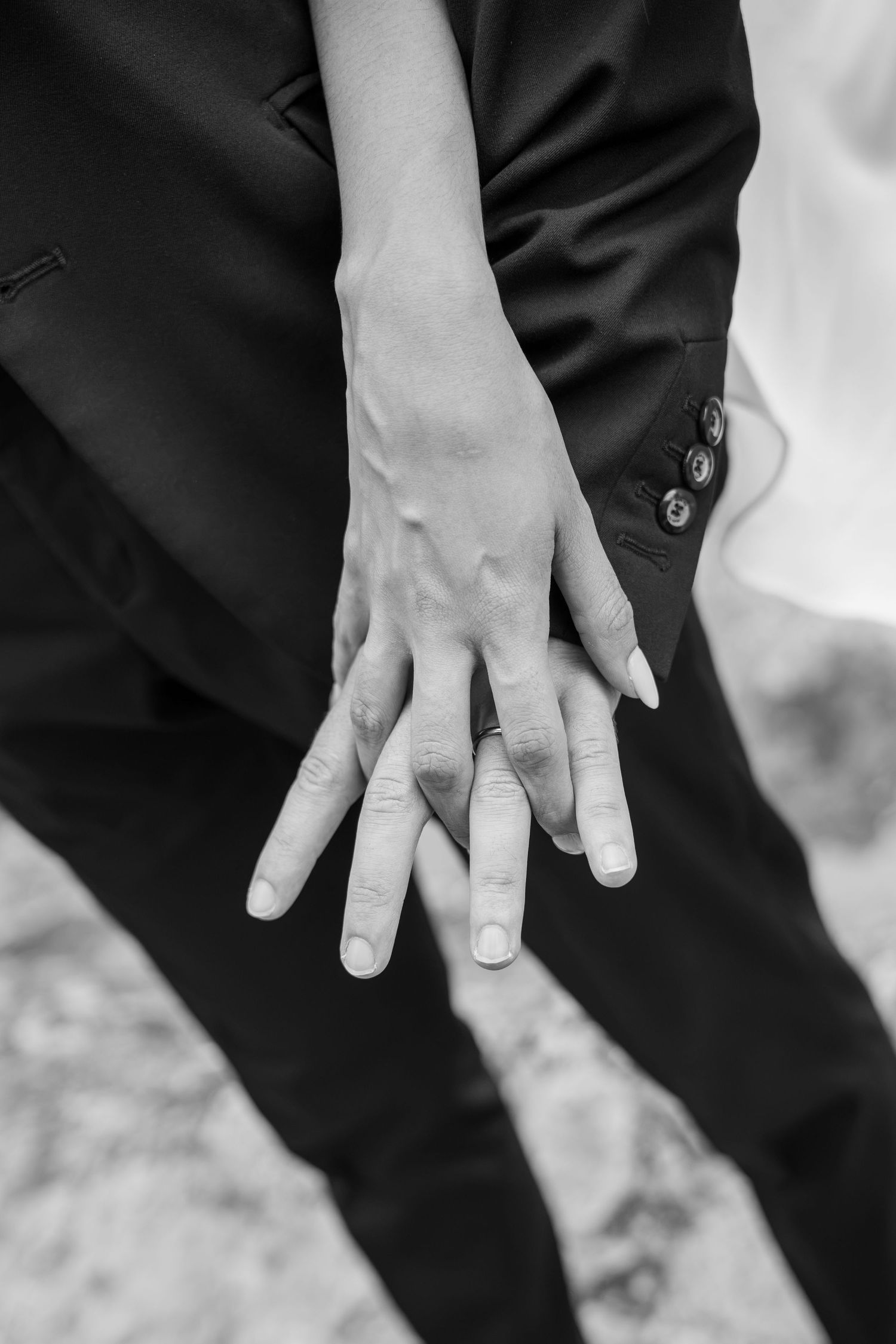 Black and white photo of two hands overlapping behind a person's back, one wearing a ring, against a dark suit.