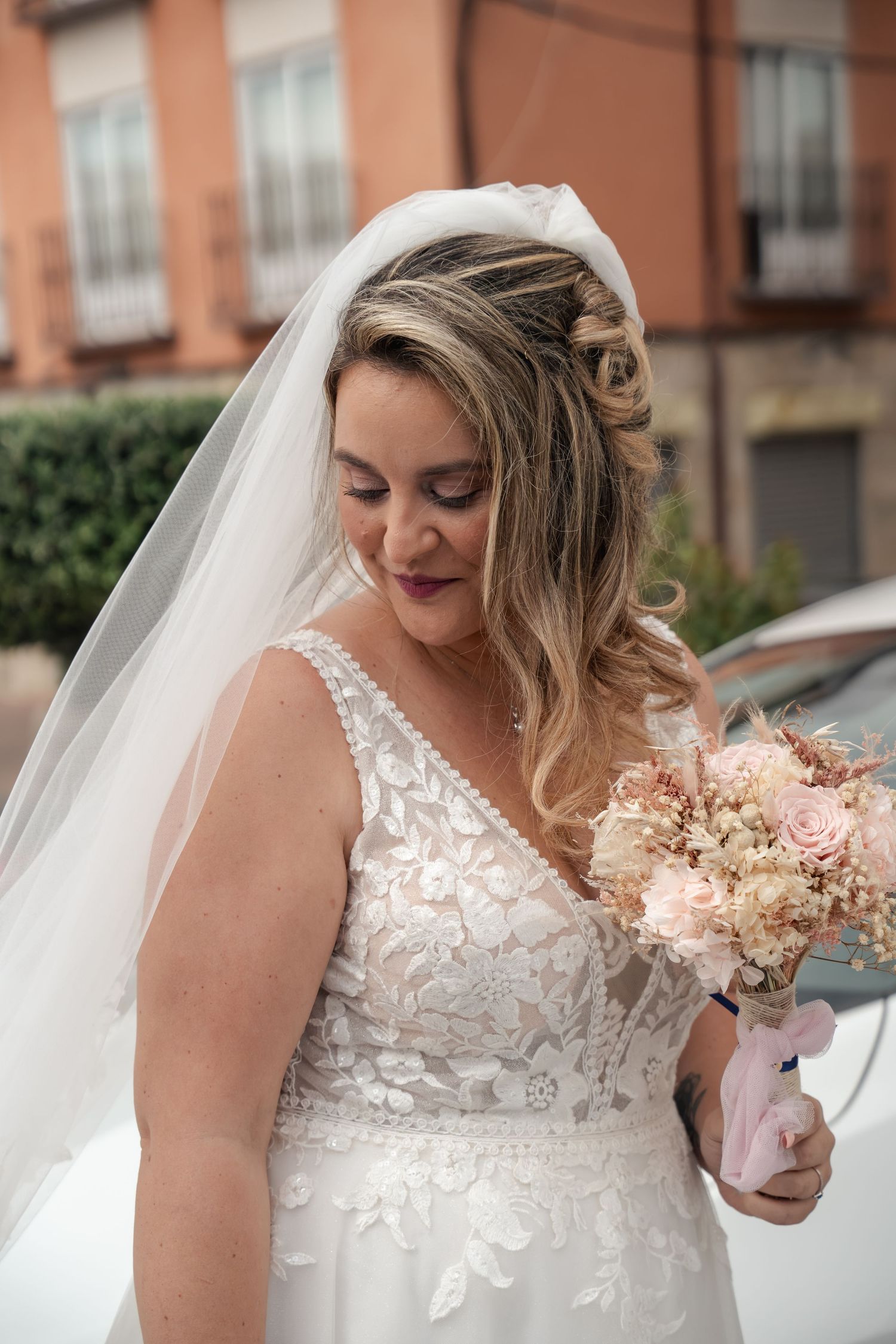 Smiling bride in a lace wedding gown and long veil looks down at her pink and blush rose bouquet while standing outdoors.