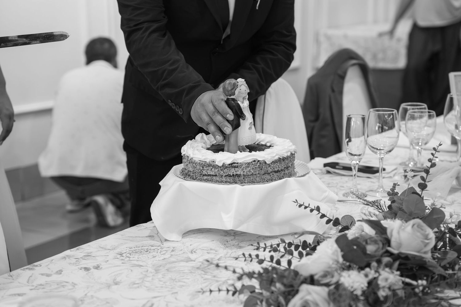 Black and white photo of a man in a suit cutting a wedding cake on a draped table with flowers and wine glasses at a reception.