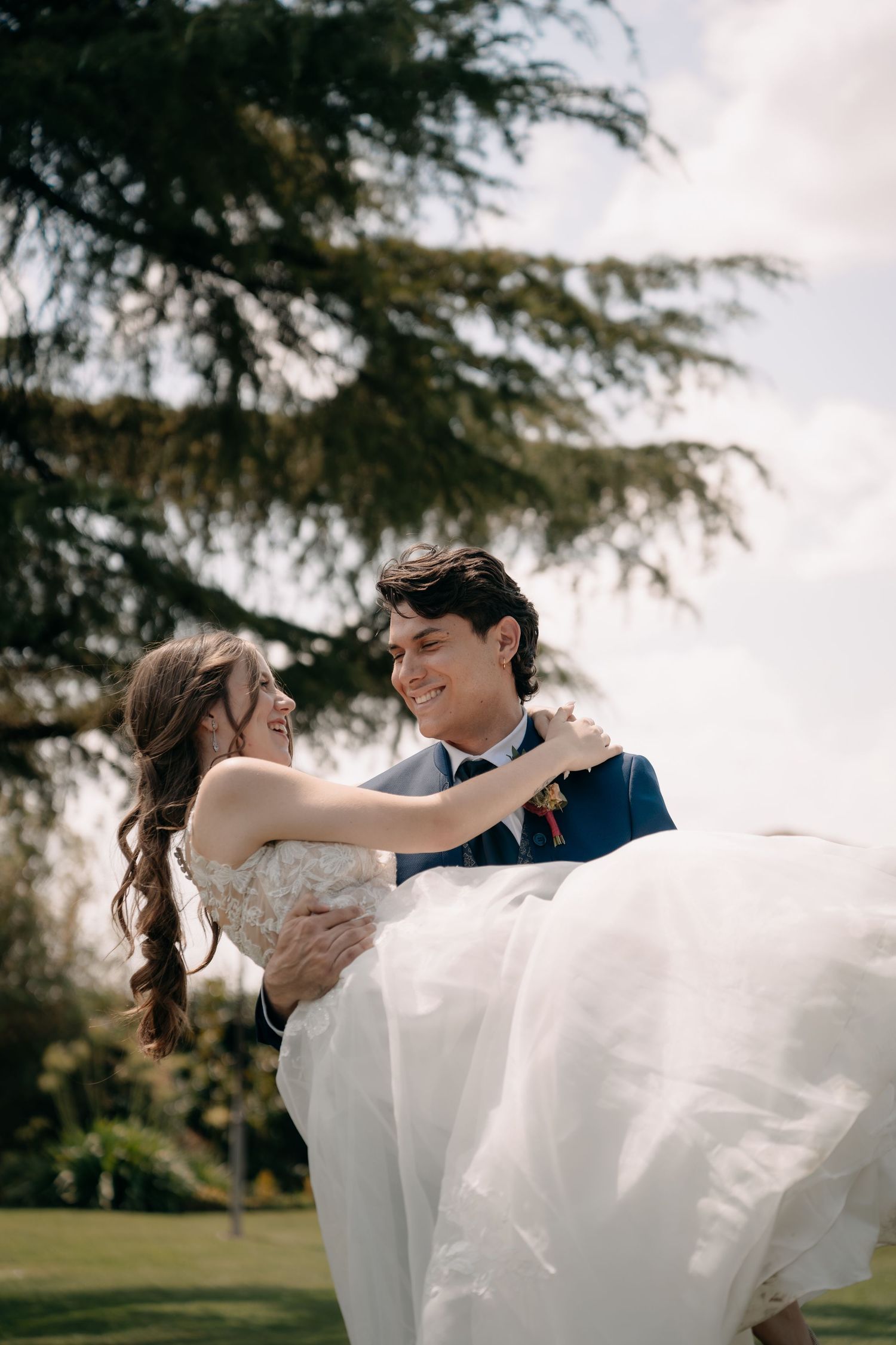 Smiling groom carries laughing bride in white dress outdoors with a large pine tree behind them.