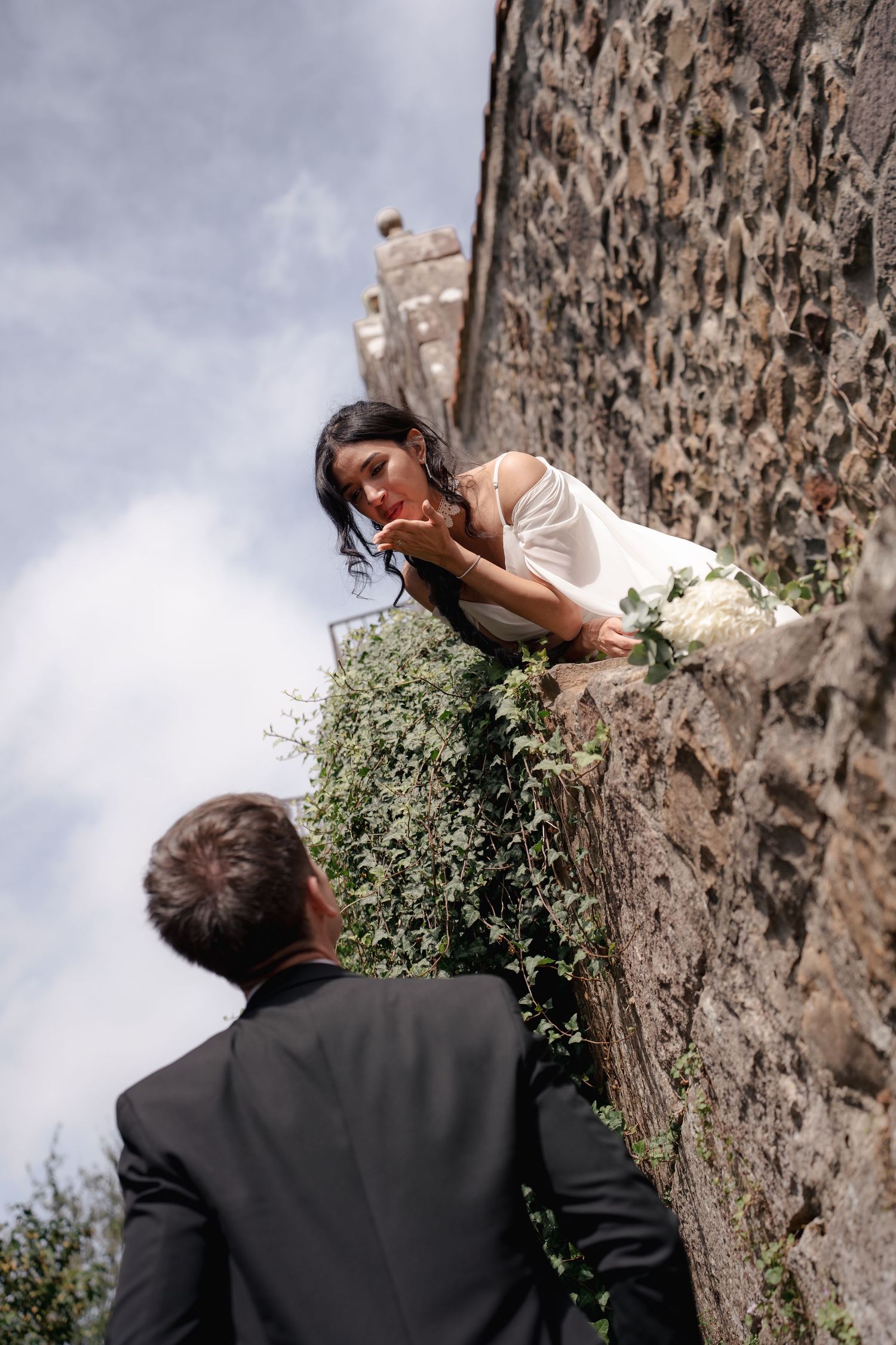 Bride in white dress leans over ivy-covered stone wall gazing down at groom in black suit during outdoor wedding.