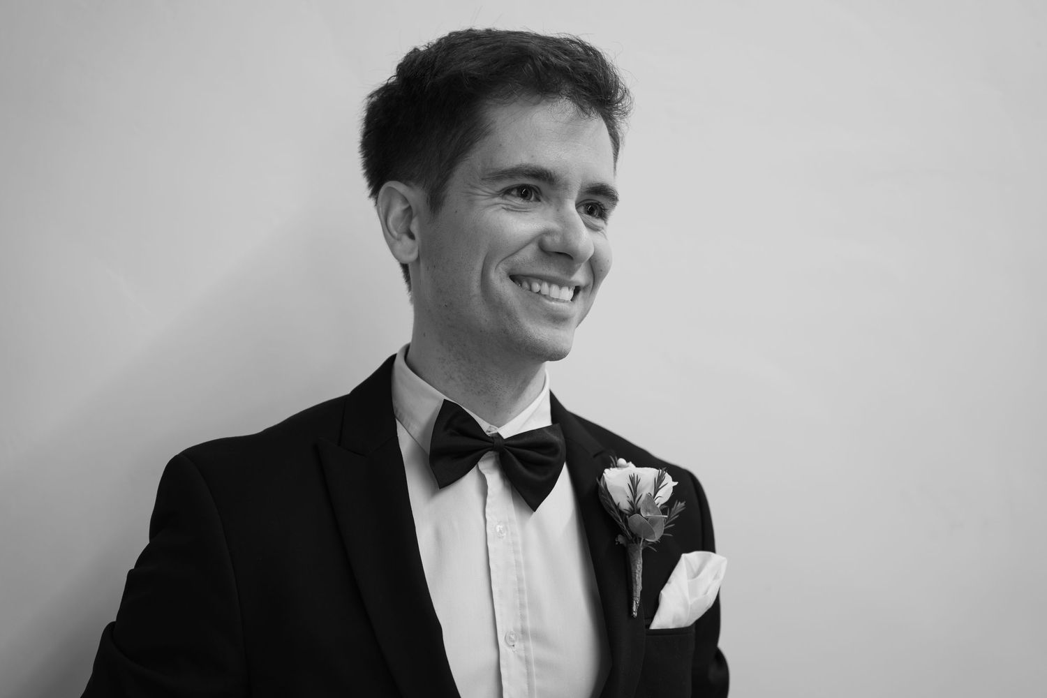 Smiling man in a black tuxedo, bow tie and floral boutonniere with a pocket square, posed in a black and white portrait.
