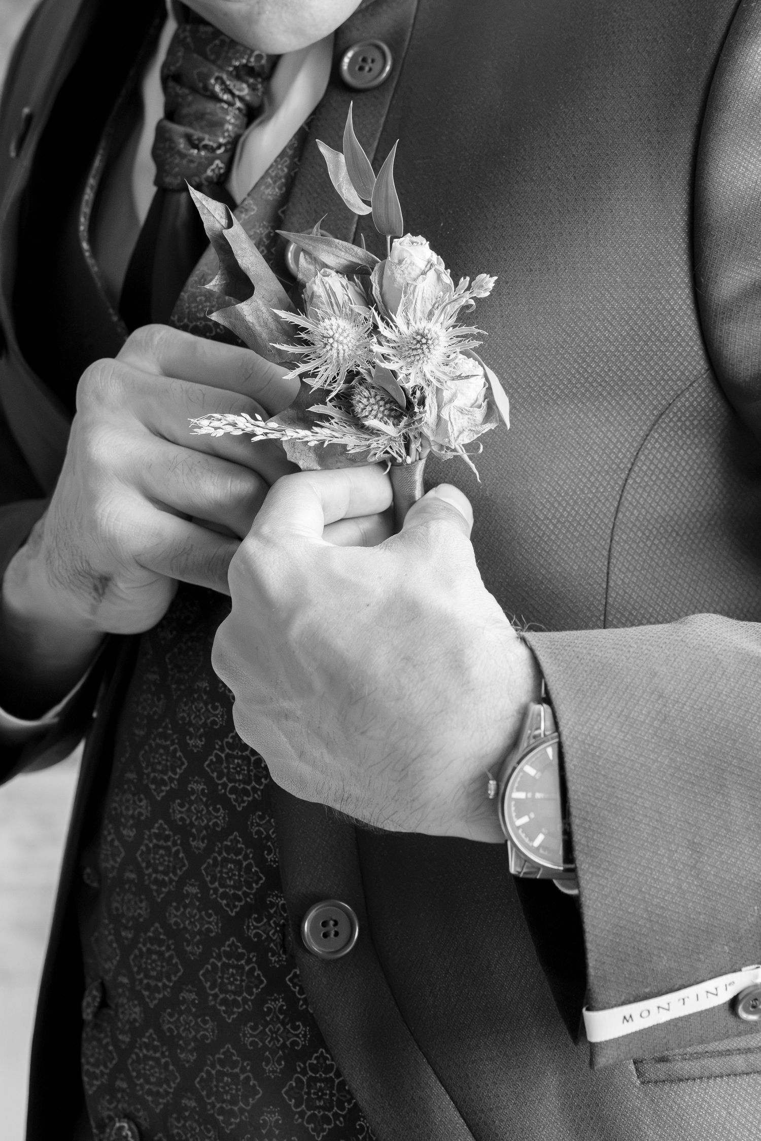 Black and white photo of a groom pinning a floral boutonniere on his suit jacket, wearing a watch.