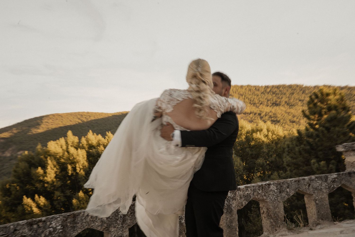 Bride and groom embracing on stone terrace with scenic mountain forest landscape in background.