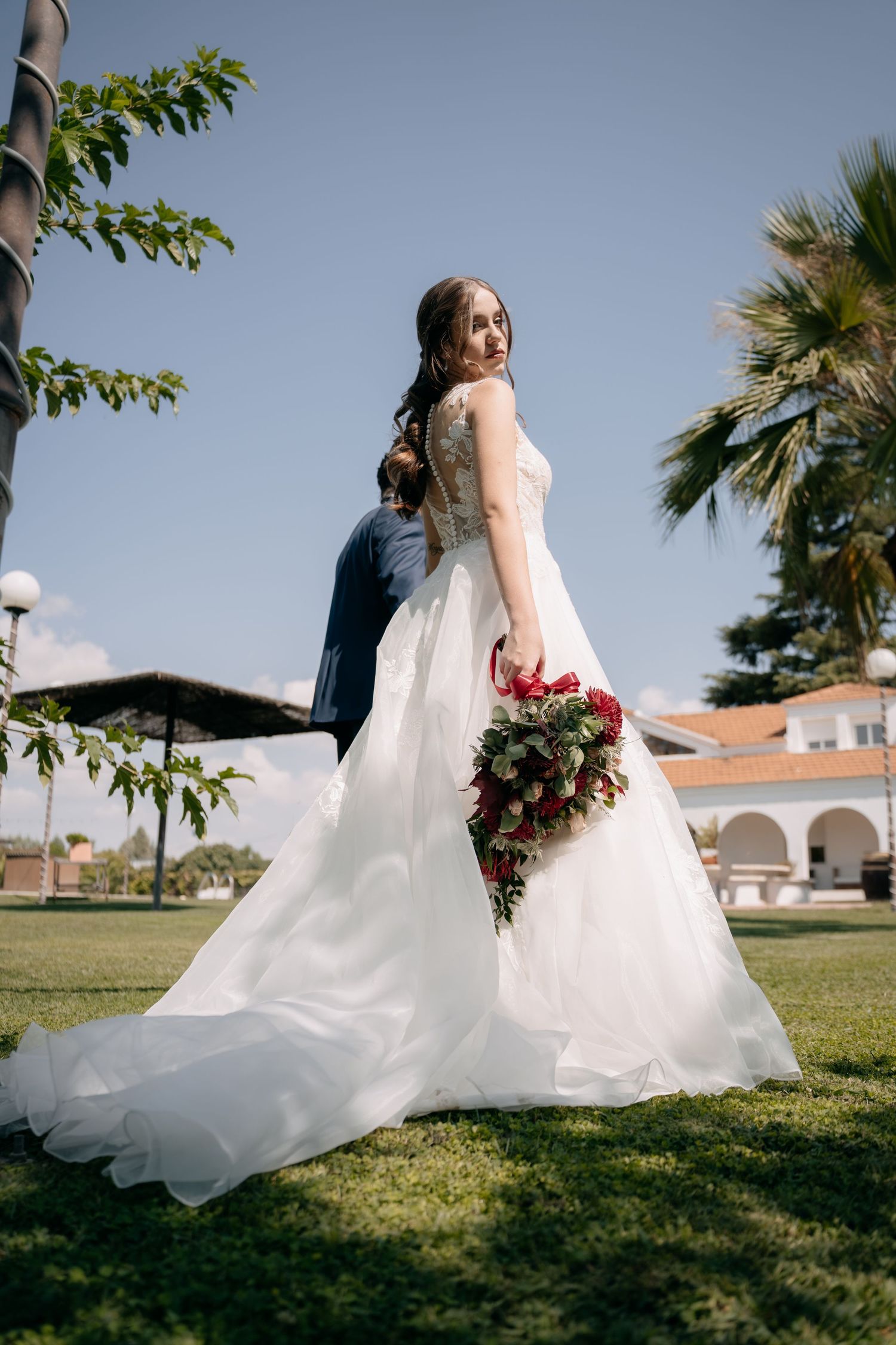 Elegant bride in full white gown holding red bouquet with groom embracing her in garden.