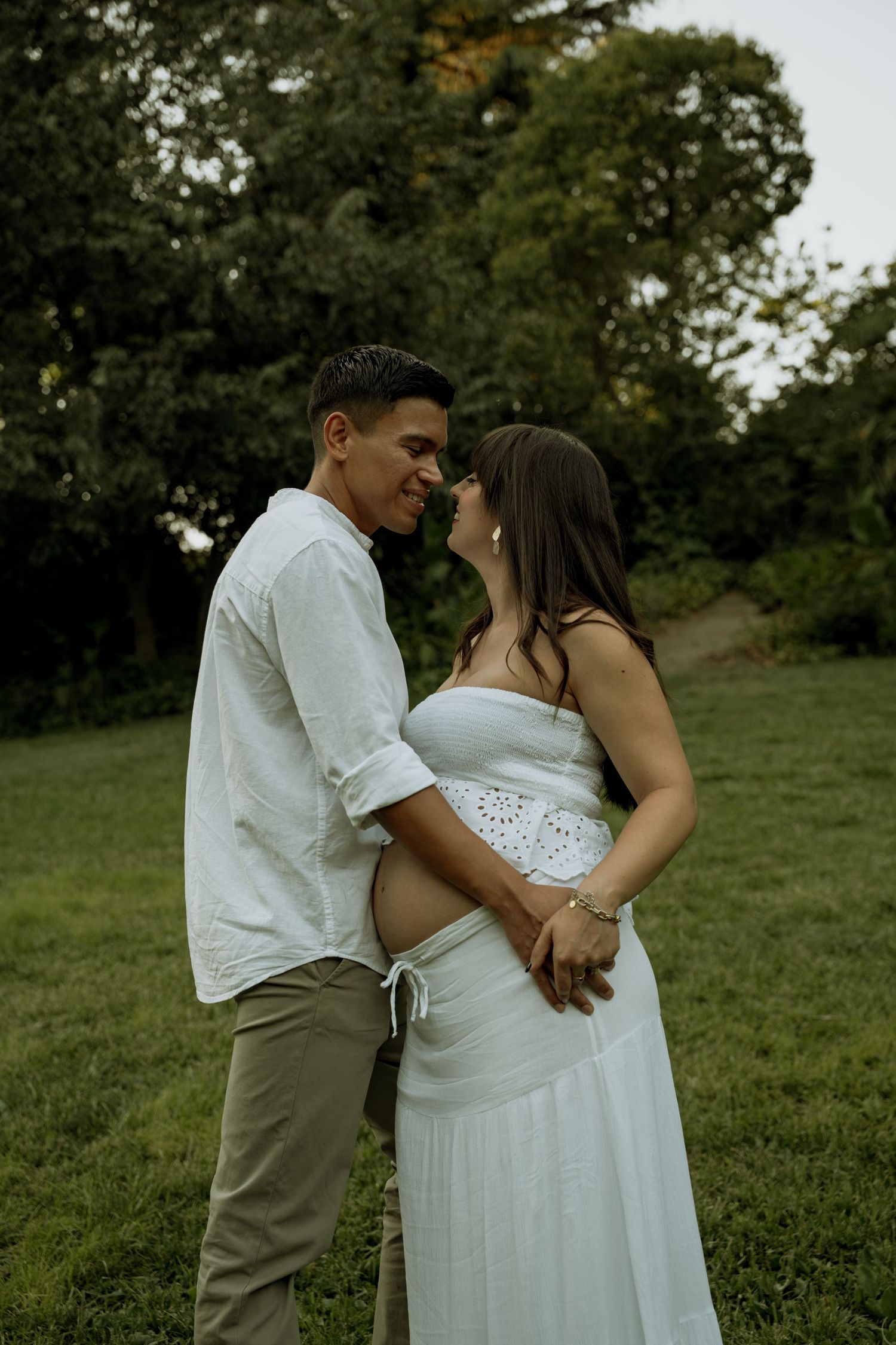 Happy expecting couple nearly kissing while embracing on a green lawn during an outdoor maternity photo session.