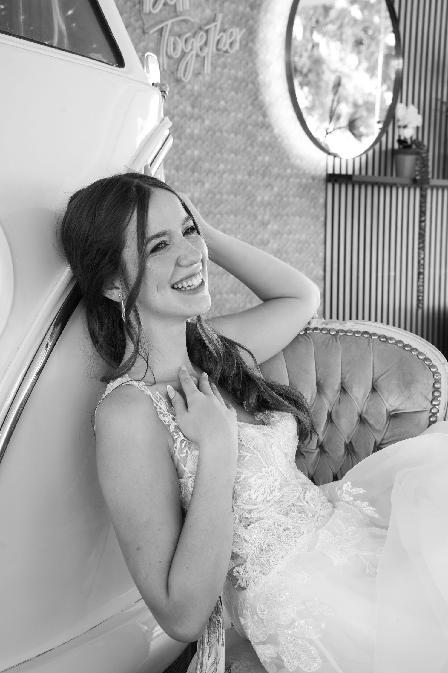 Smiling bride in a lace wedding gown leans against a vintage white car beside a tufted velvet sofa in a black and white photo.