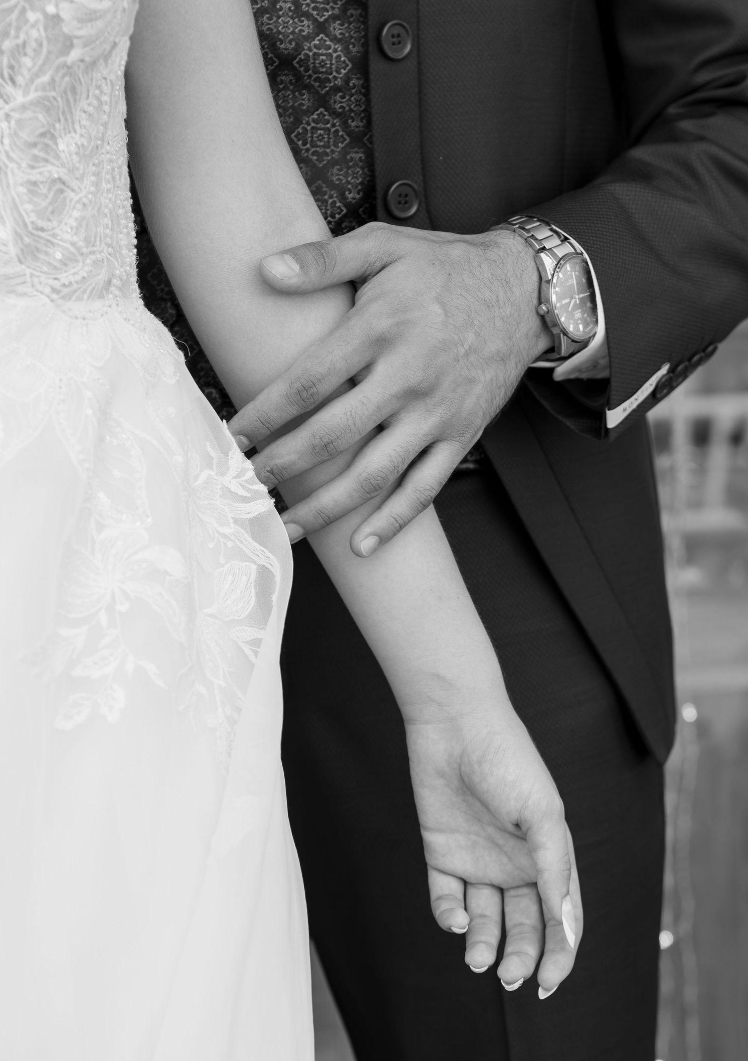 Black and white close-up of a groom's hand resting gently on a bride's forearm, with her lace wedding dress and his suit visible.