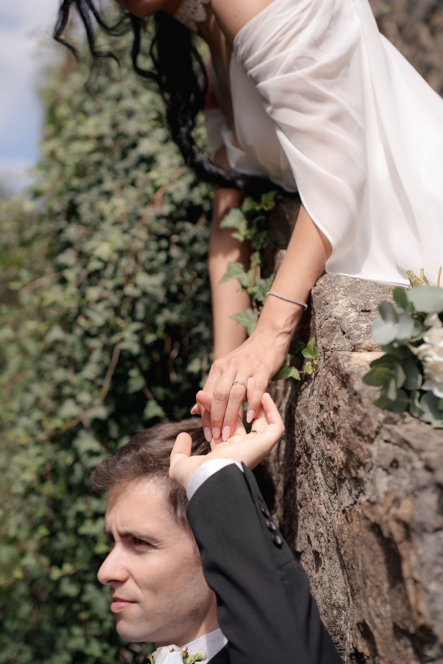 Bride in a white dress leans over an ivy-covered stone wall to hold hands with a groom in a dark suit below.