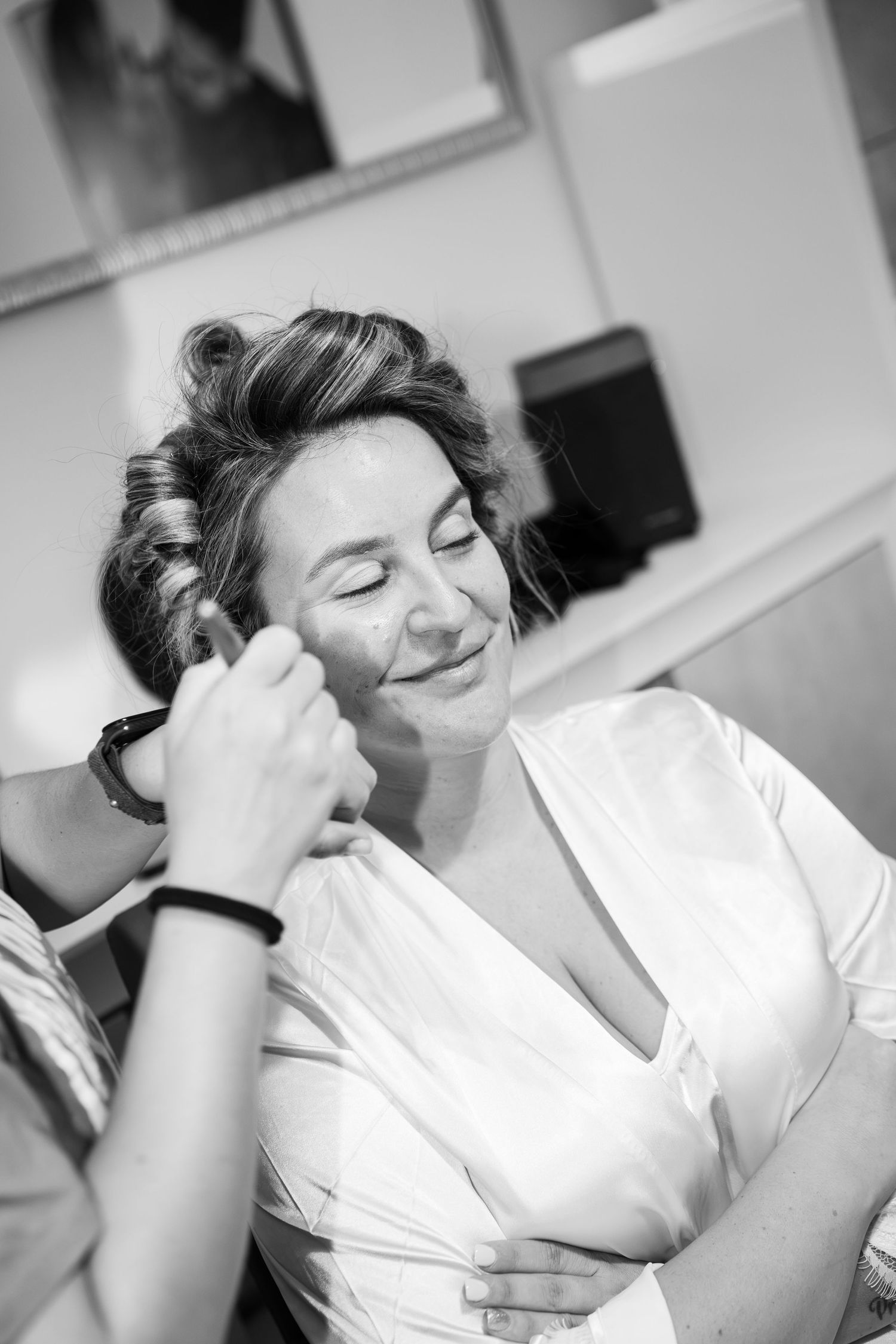 Smiling woman in a white satin robe with hair in curlers has makeup applied by a makeup artist during bridal preparation.