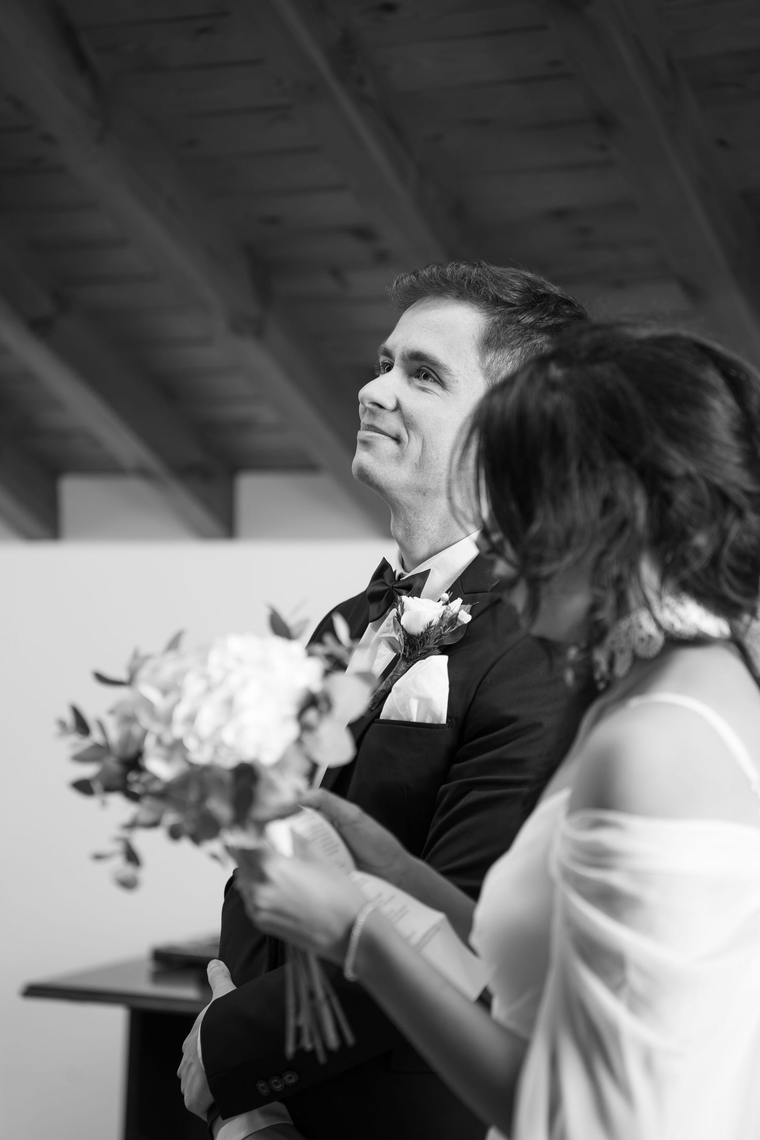 Black and white photo of a smiling groom in a tuxedo holding a bouquet with his bride during a wedding ceremony.
