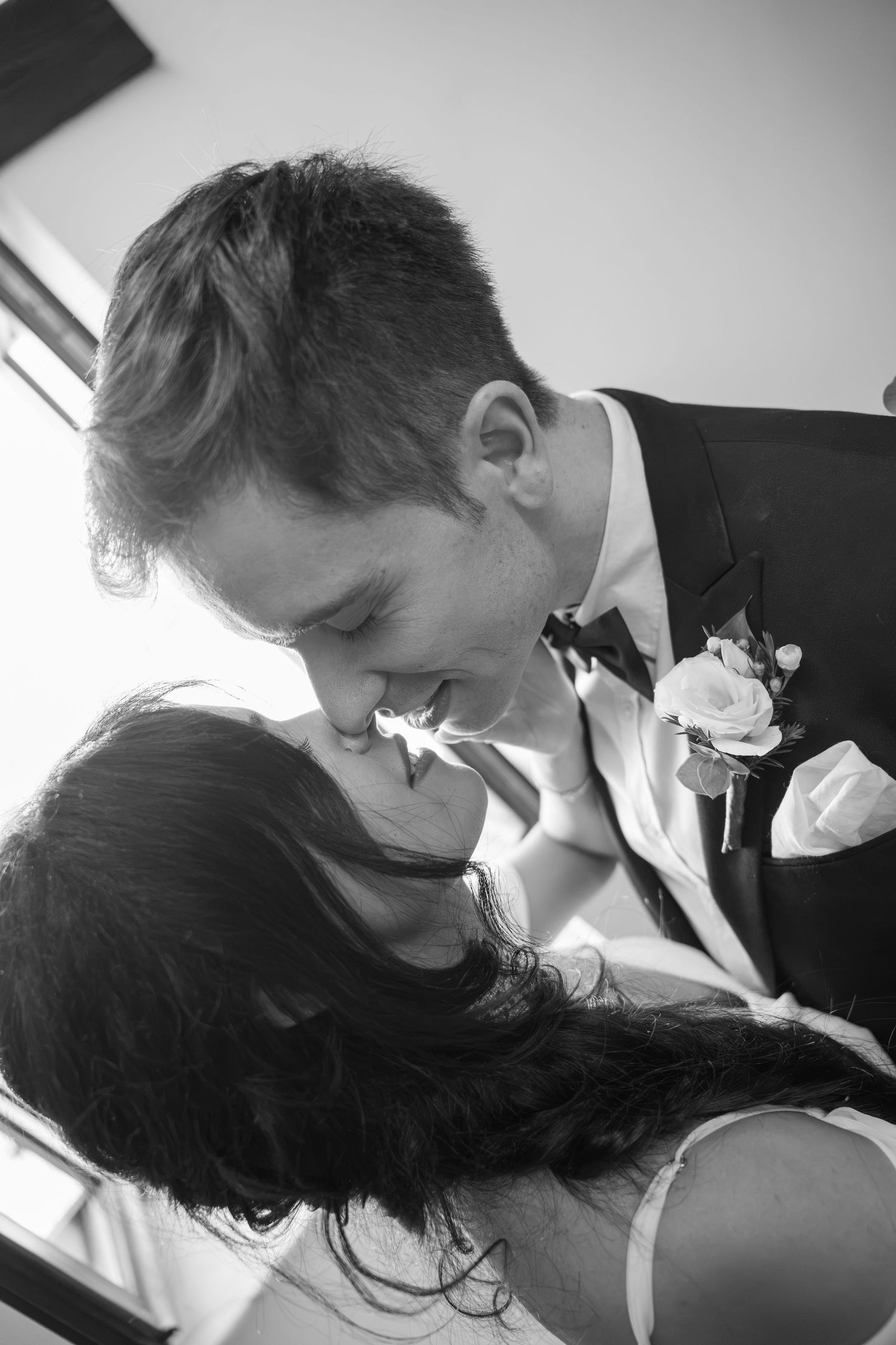 Black and white wedding photo of a groom in a suit and boutonniere leaning in to kiss his smiling bride with long dark hair.