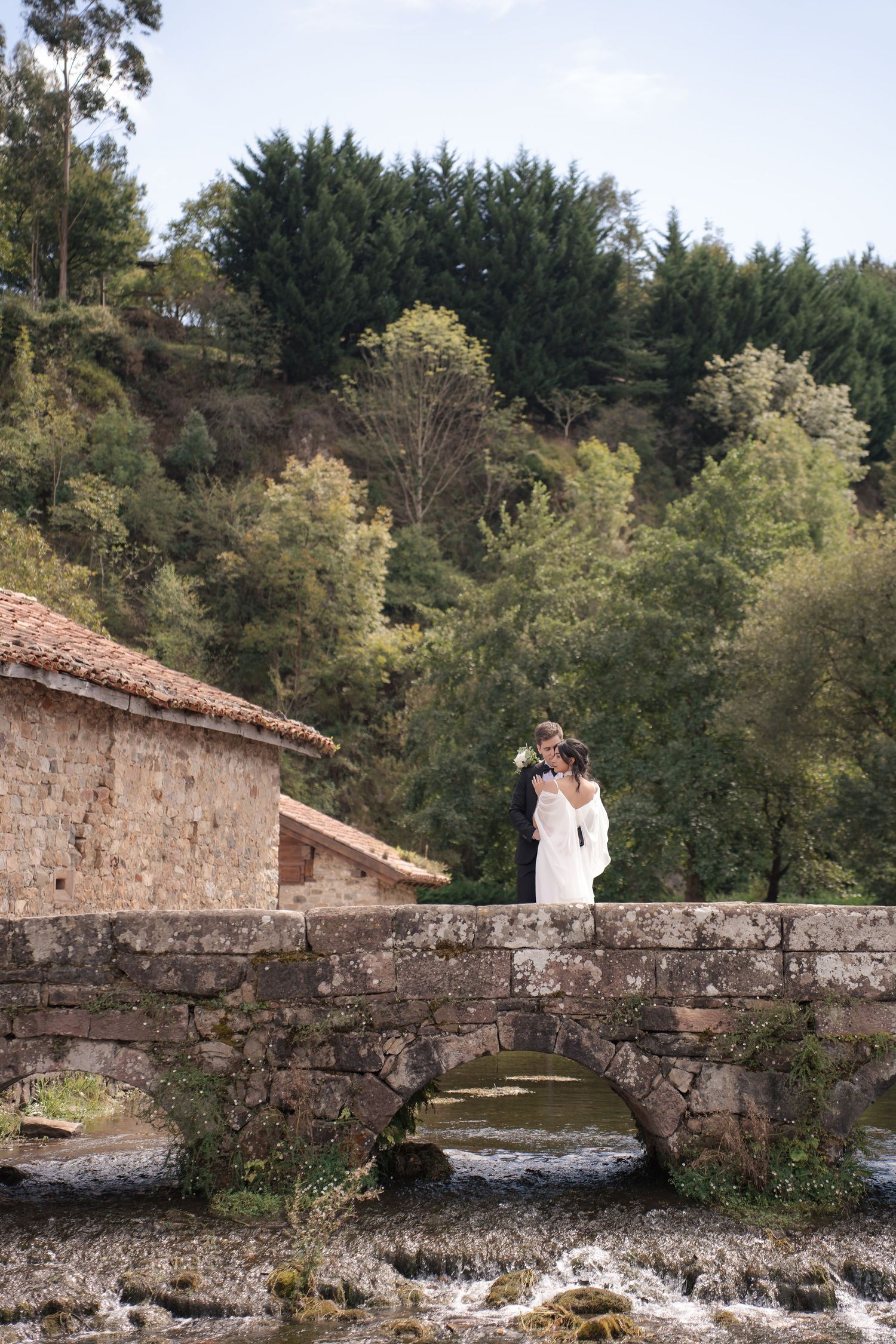 Bride and groom embrace on an old stone bridge over a rushing stream, with rustic buildings and lush green hillside trees behind them.