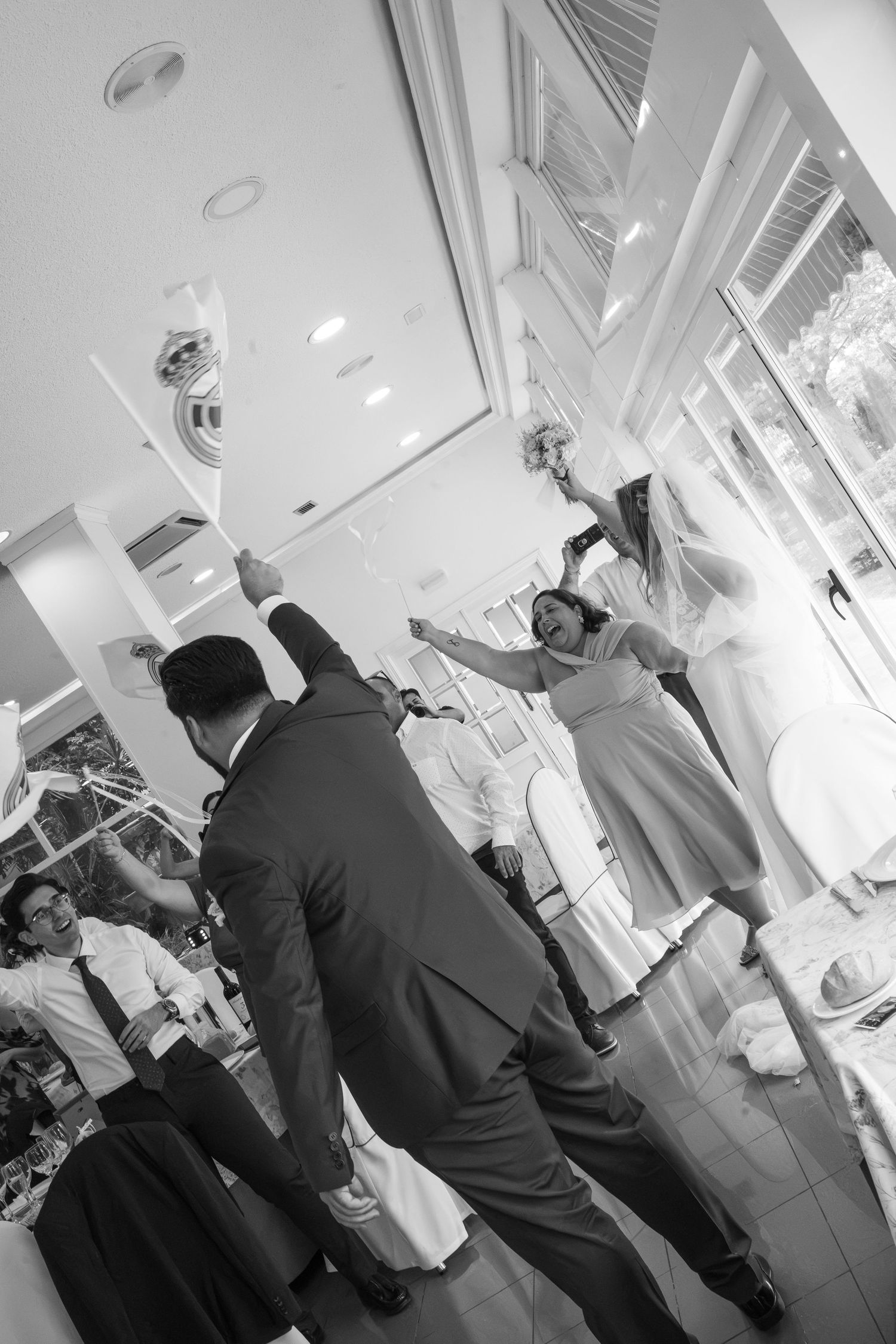 Black and white photo of wedding guests cheering and raising their arms as a bride holding a bouquet dances in a bright reception hall.