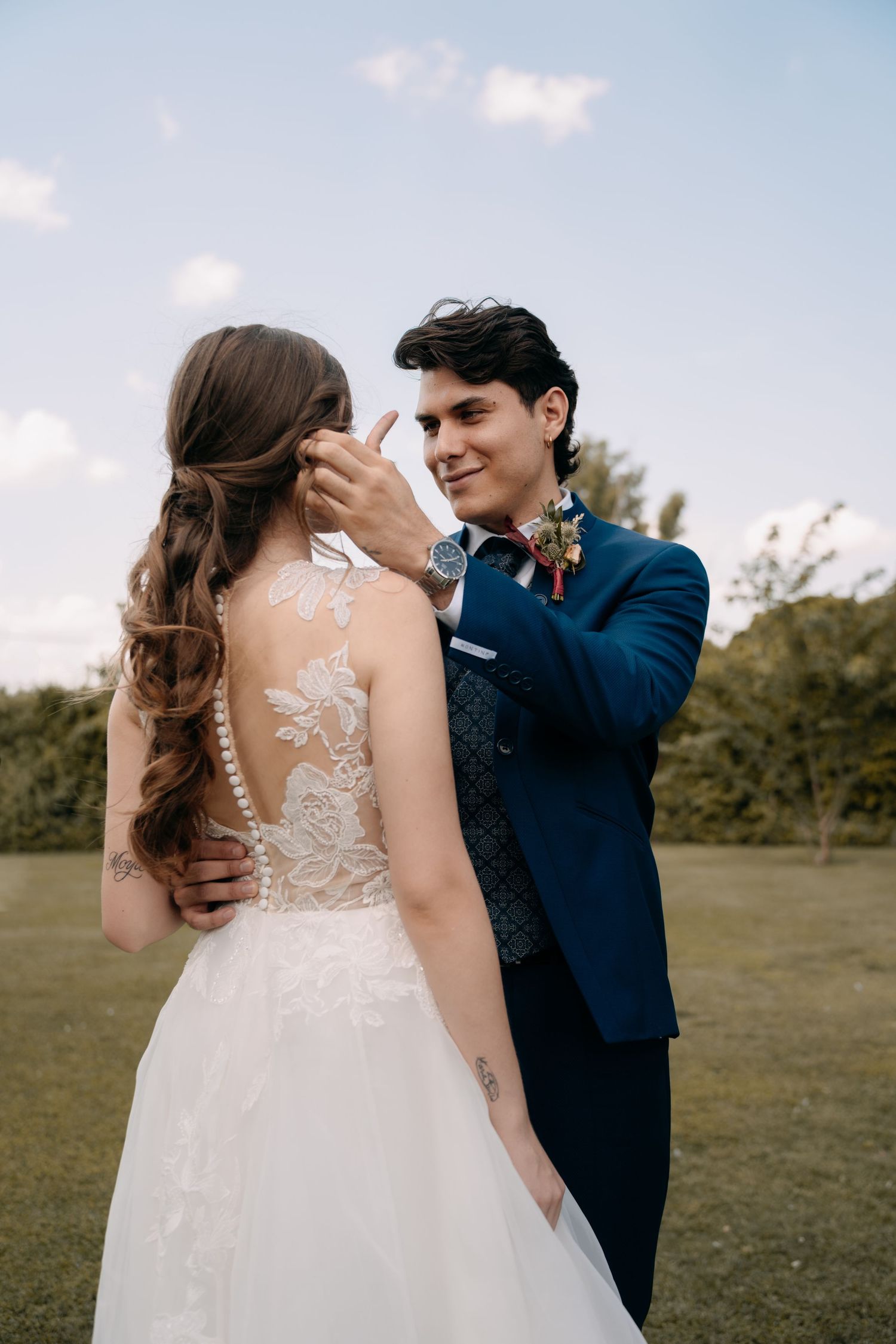 Smiling groom in a blue suit with floral boutonniere gently tucks hair behind the ear of a bride in a lace wedding gown outdoors.