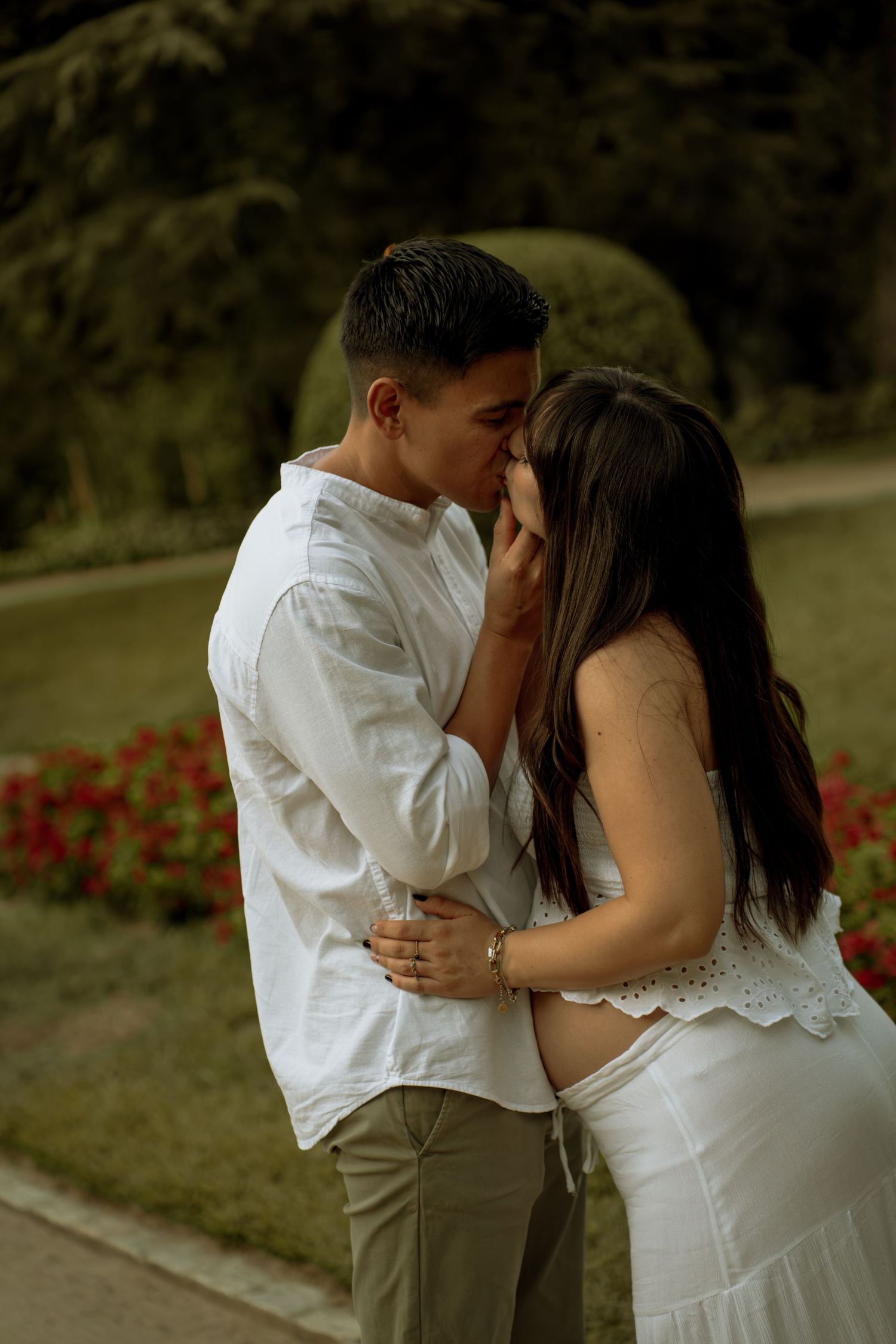 Expecting couple sharing an intimate kiss in a garden park, with the woman cradling her baby bump in a white dress.