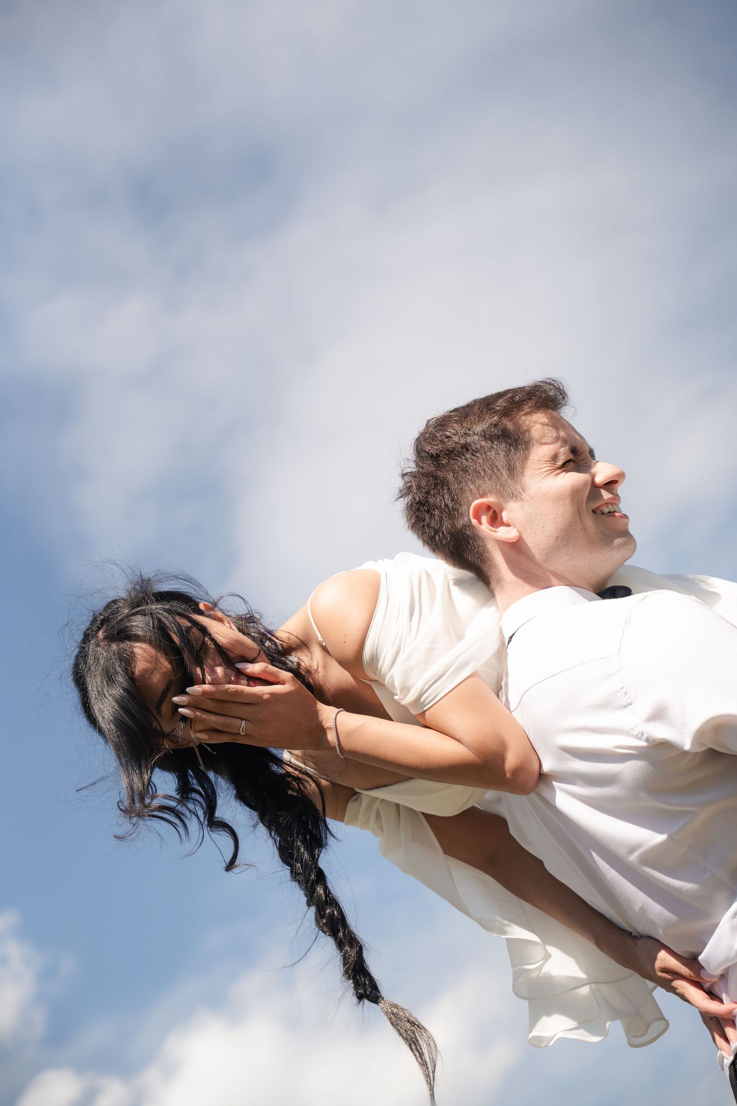 A joyful couple embraces outdoors, the man lifting the woman against a bright blue cloudy sky.
