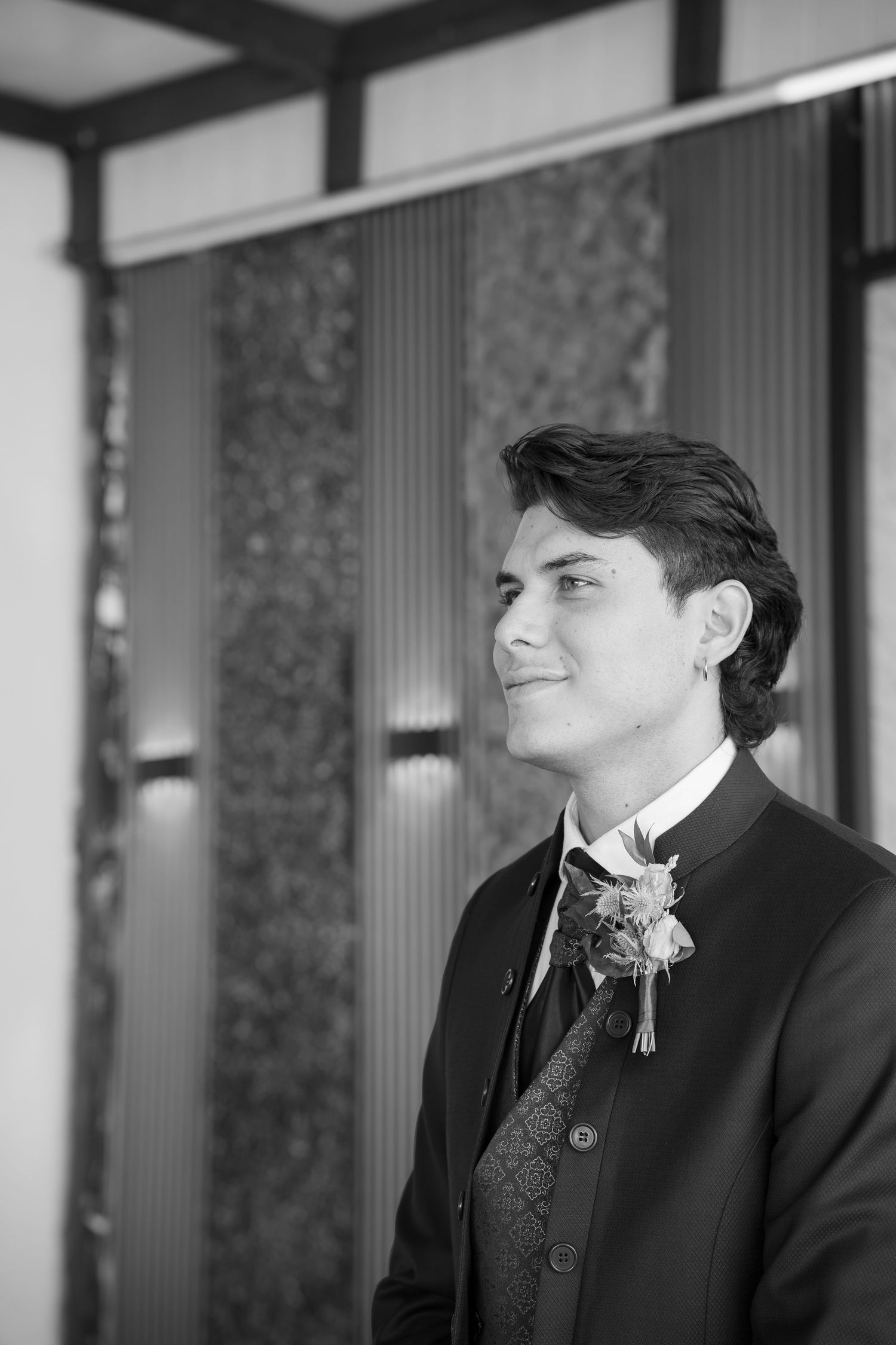 Black and white portrait of a smiling groom in a dark vest and jacket with a floral boutonniere, standing indoors before a textured wall.