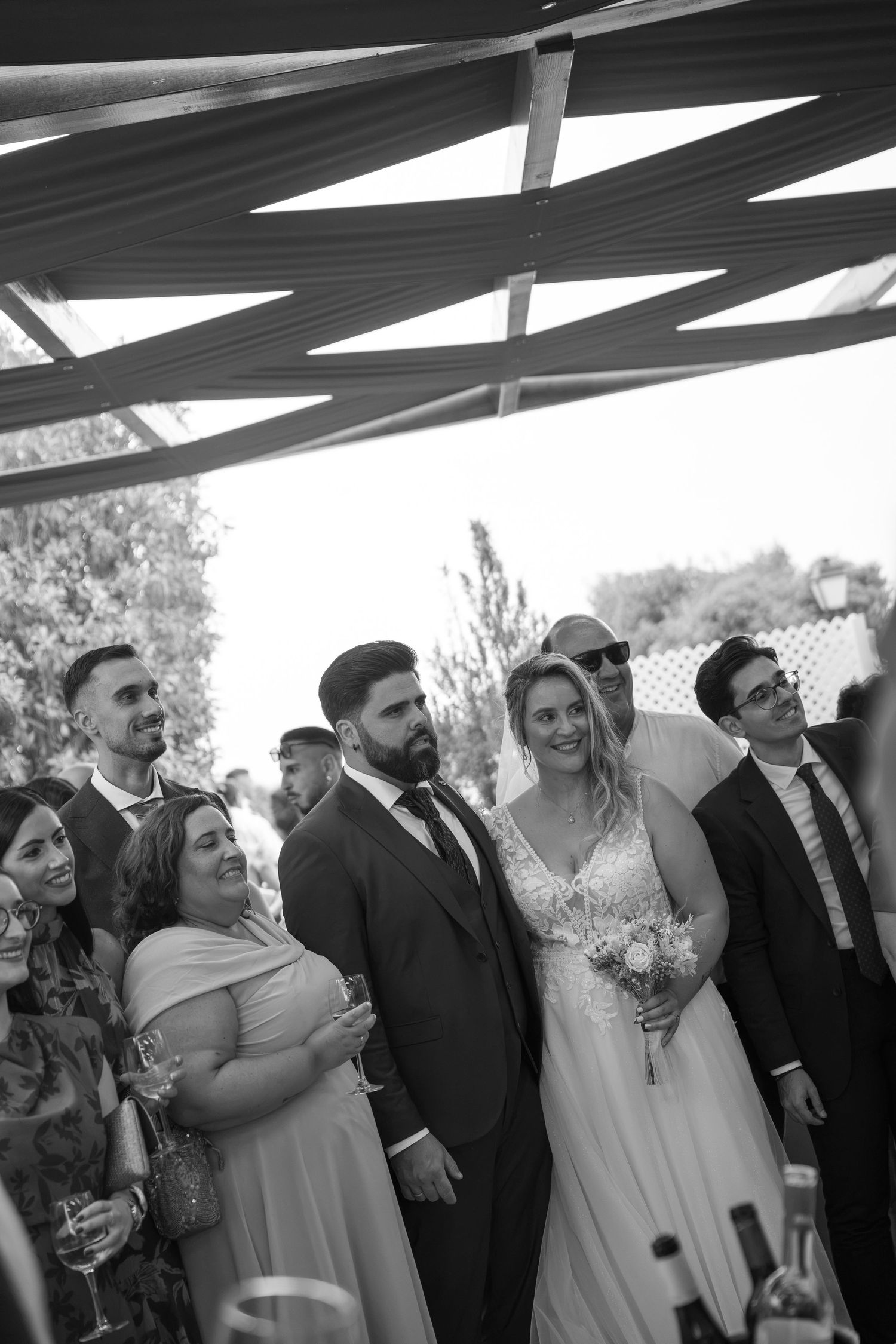 Black and white photo of a bride holding a bouquet beside her groom, with smiling wedding guests raising glasses under a geometric canopy.