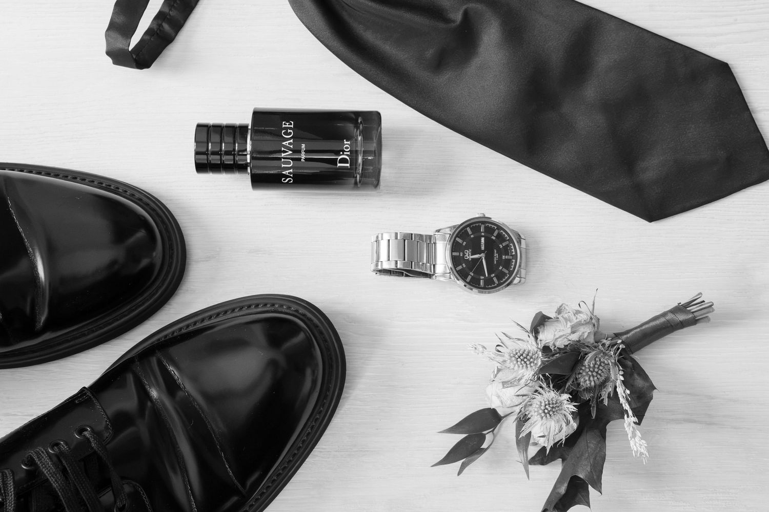 Black and white flat lay of polished leather dress shoes, a silk tie, a Dior Sauvage cologne bottle, a steel wristwatch and a boutonniere.