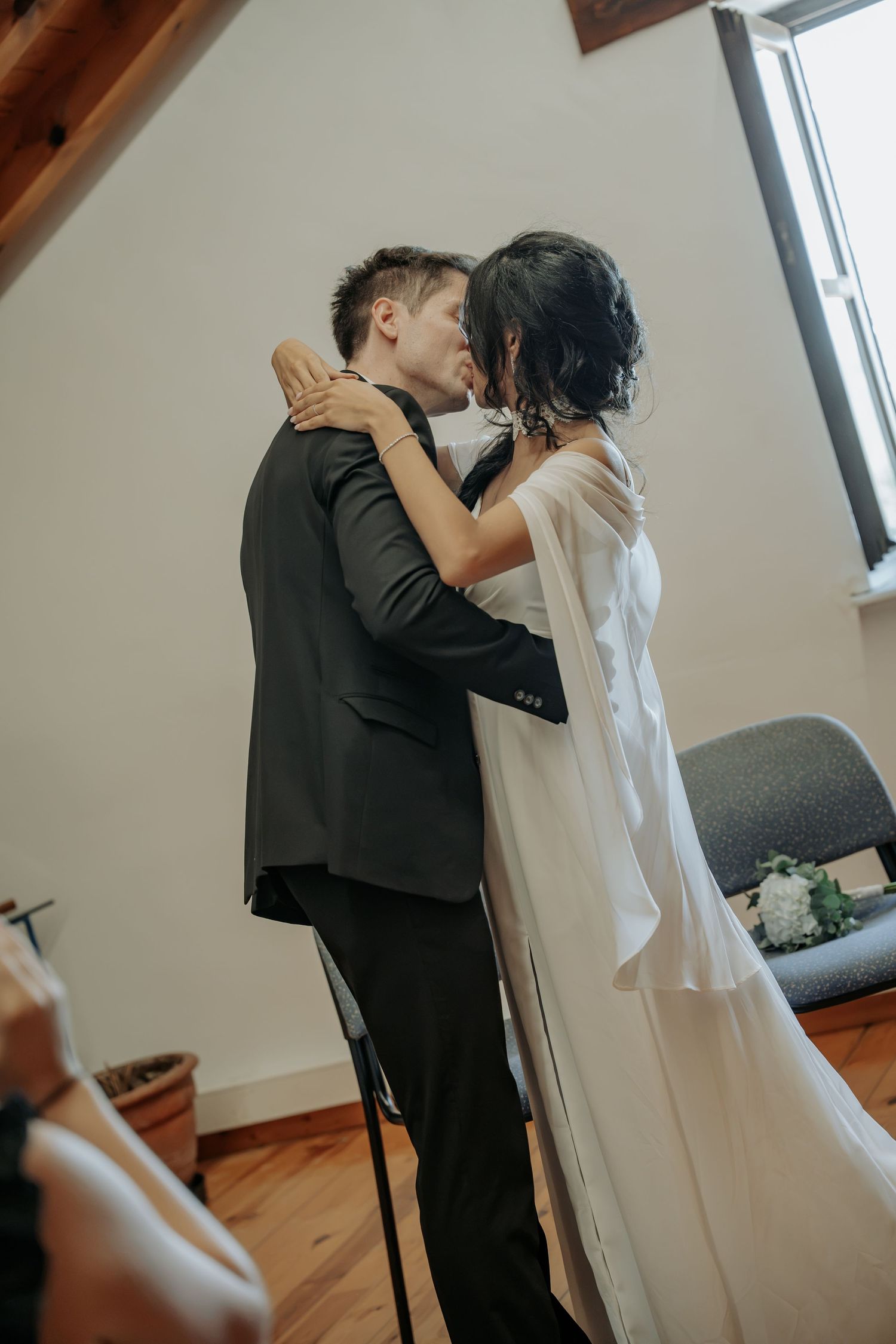 Bride in a flowing white gown and groom in a dark suit share a kiss in a bright room with wooden floors.