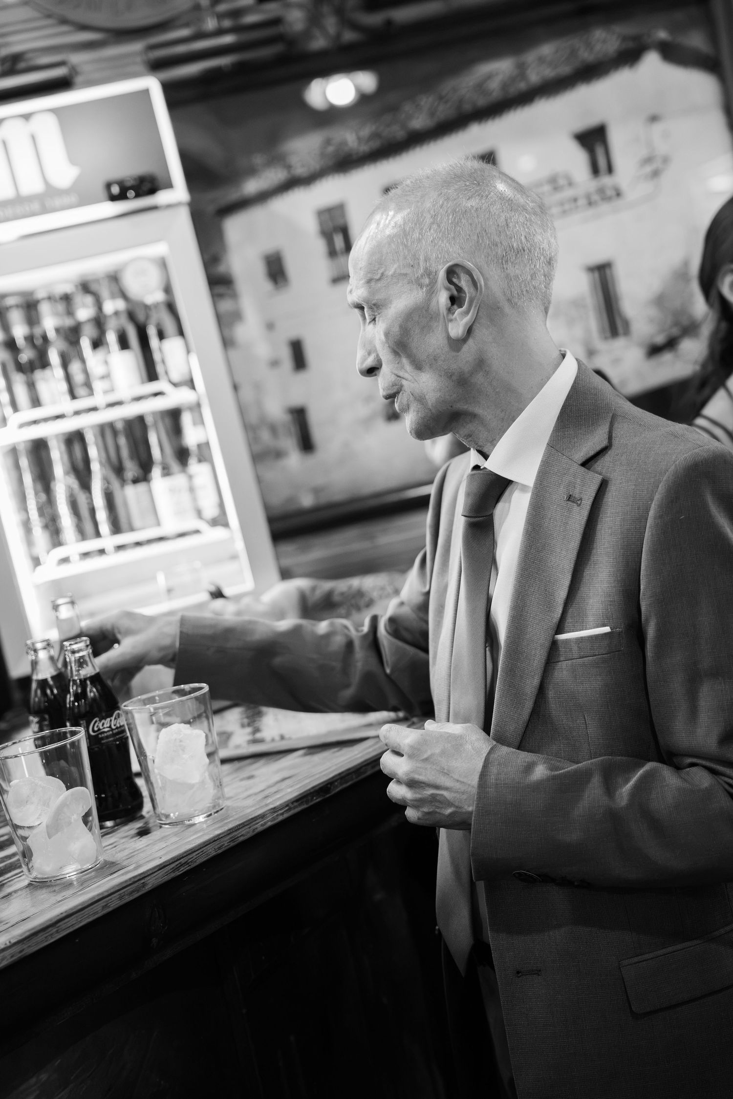 Elderly man in a suit and tie stands at a rustic bar counter beside glasses of ice and a lit refrigerator of bottled drinks.