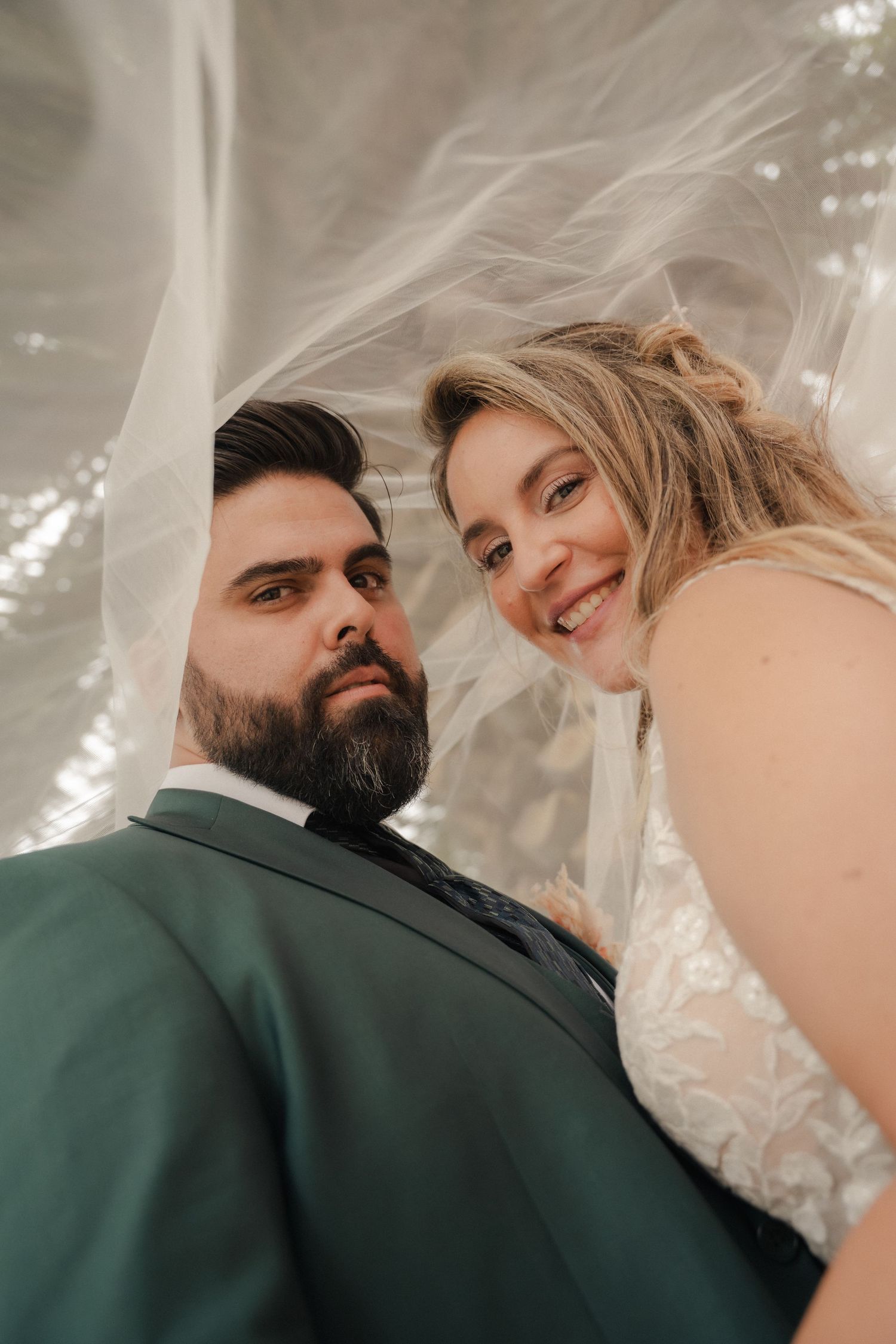 Bride in a lace gown and groom in a green suit smile for a low-angle wedding portrait beneath a flowing sheer veil.