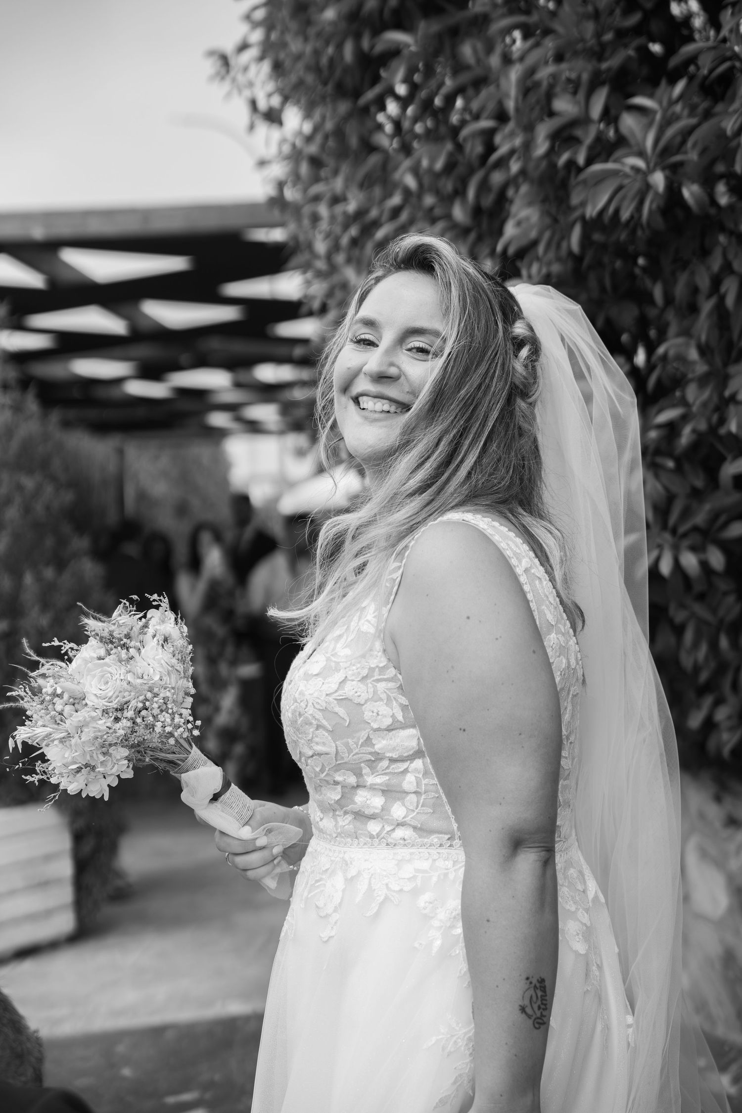 Smiling bride in a lace wedding gown and veil holds a bouquet of dried flowers at an outdoor ceremony in black and white.