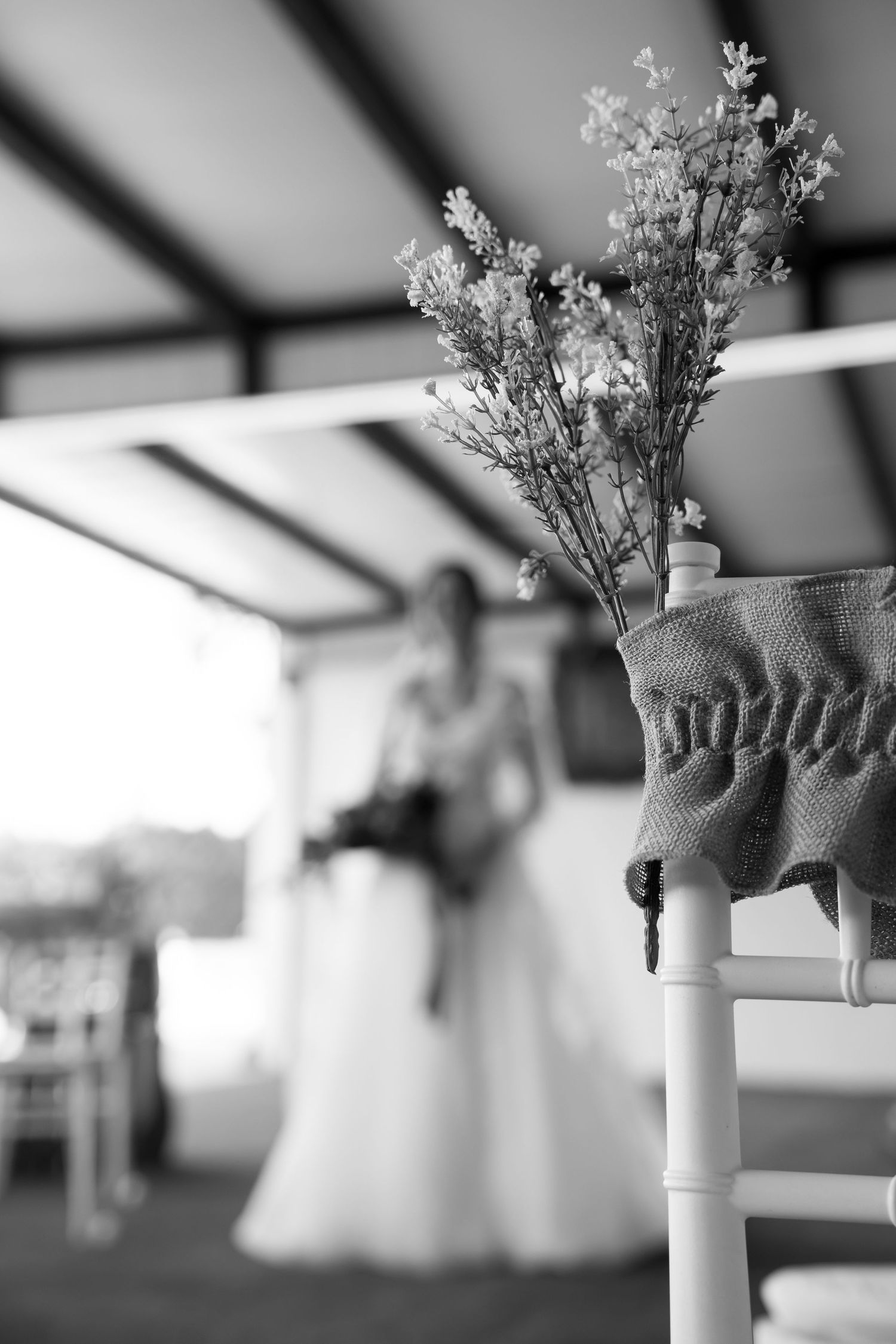 Black and white wedding photo featuring floral chair decor in foreground with blurred bride holding bouquet in background.
