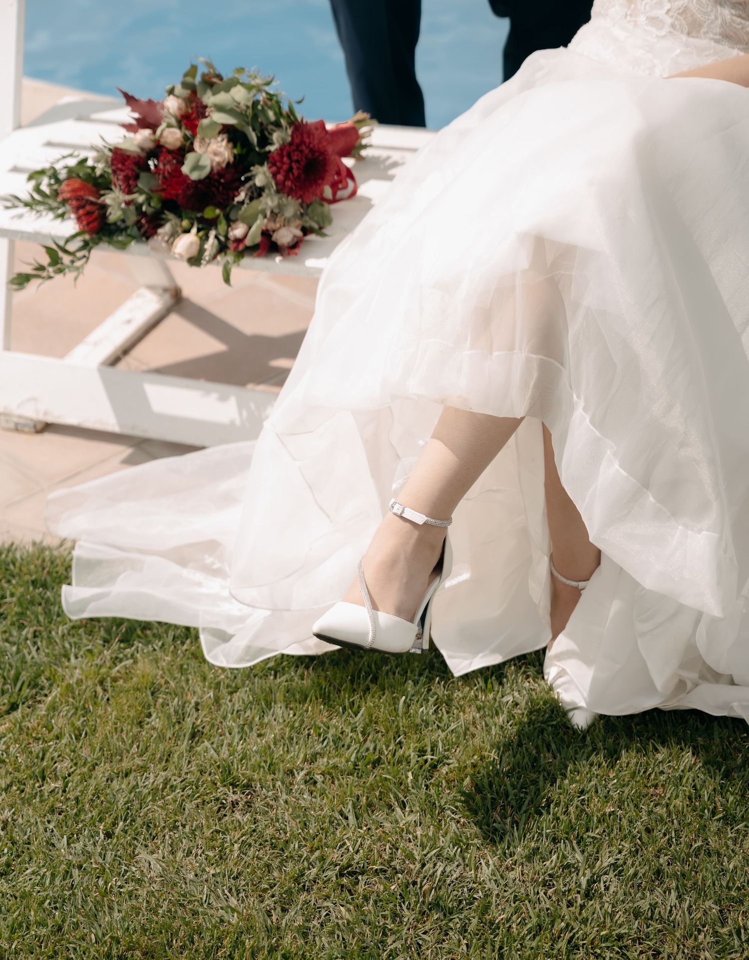 Bride in white gown with heels seated near a red floral bouquet beside a pool on grass.