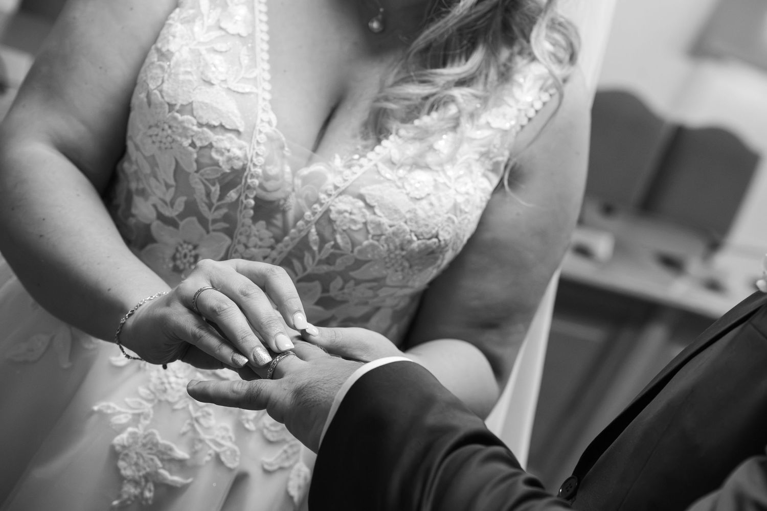 Black and white photo of a bride in a lace wedding gown placing a wedding ring on the groom's finger during the ceremony.