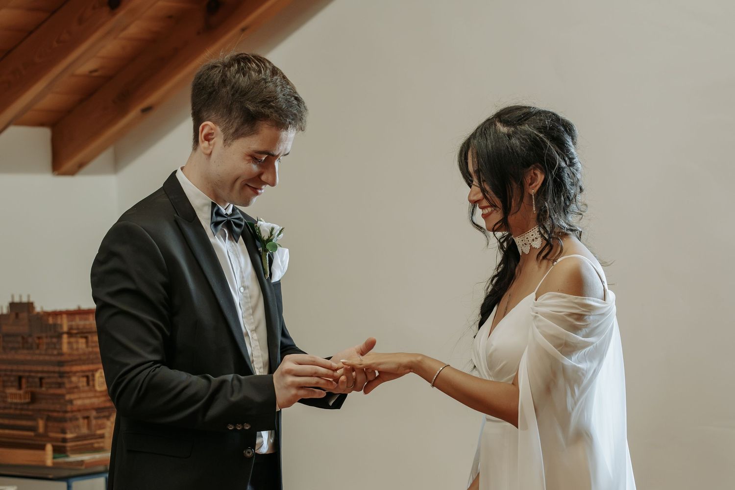 Groom in a black tuxedo and bow tie smiles as he places a wedding ring on the bride's finger during an indoor ceremony.