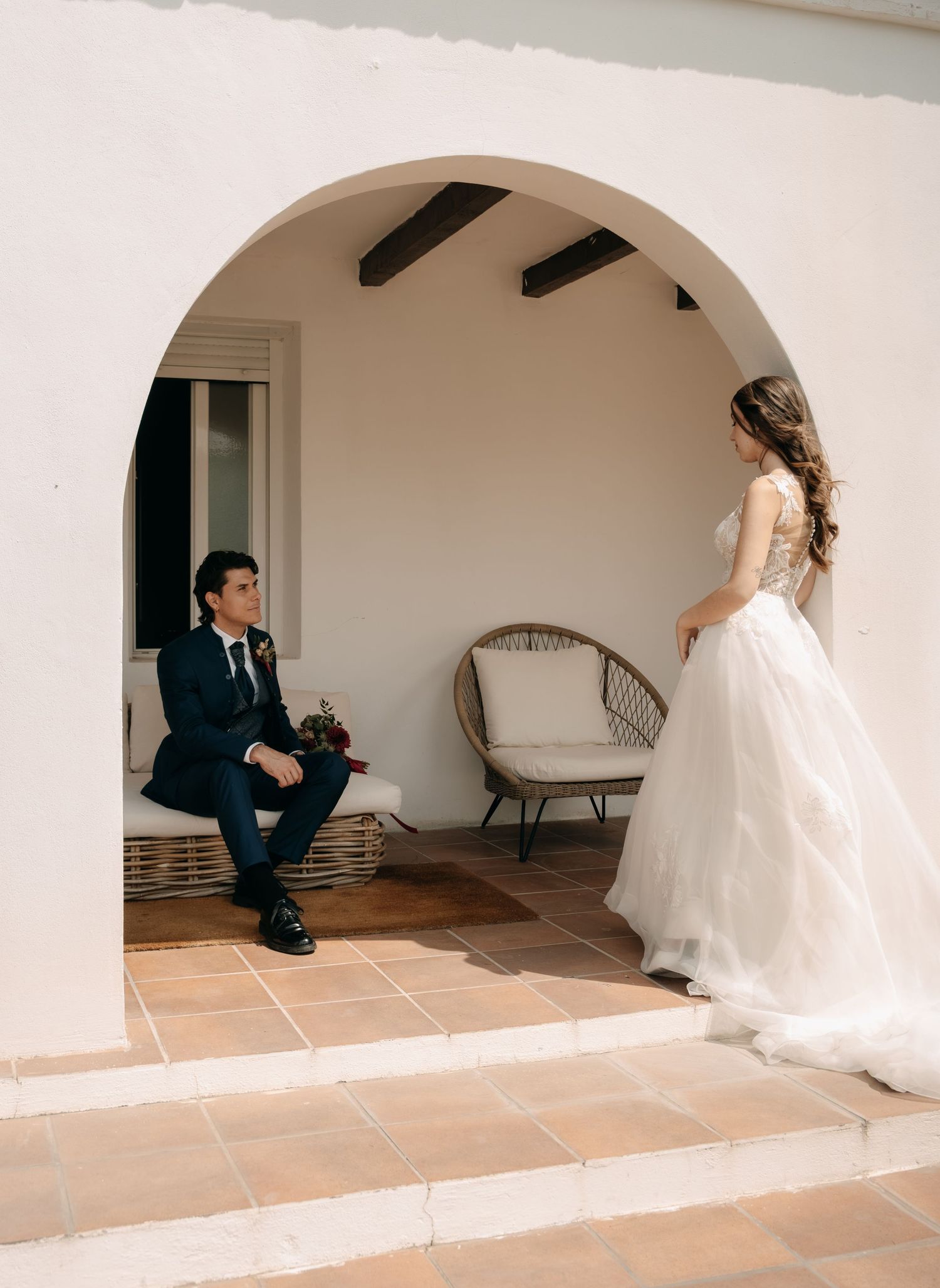 Groom in navy suit sits under white arch as bride in flowing white gown approaches during wedding first look.
