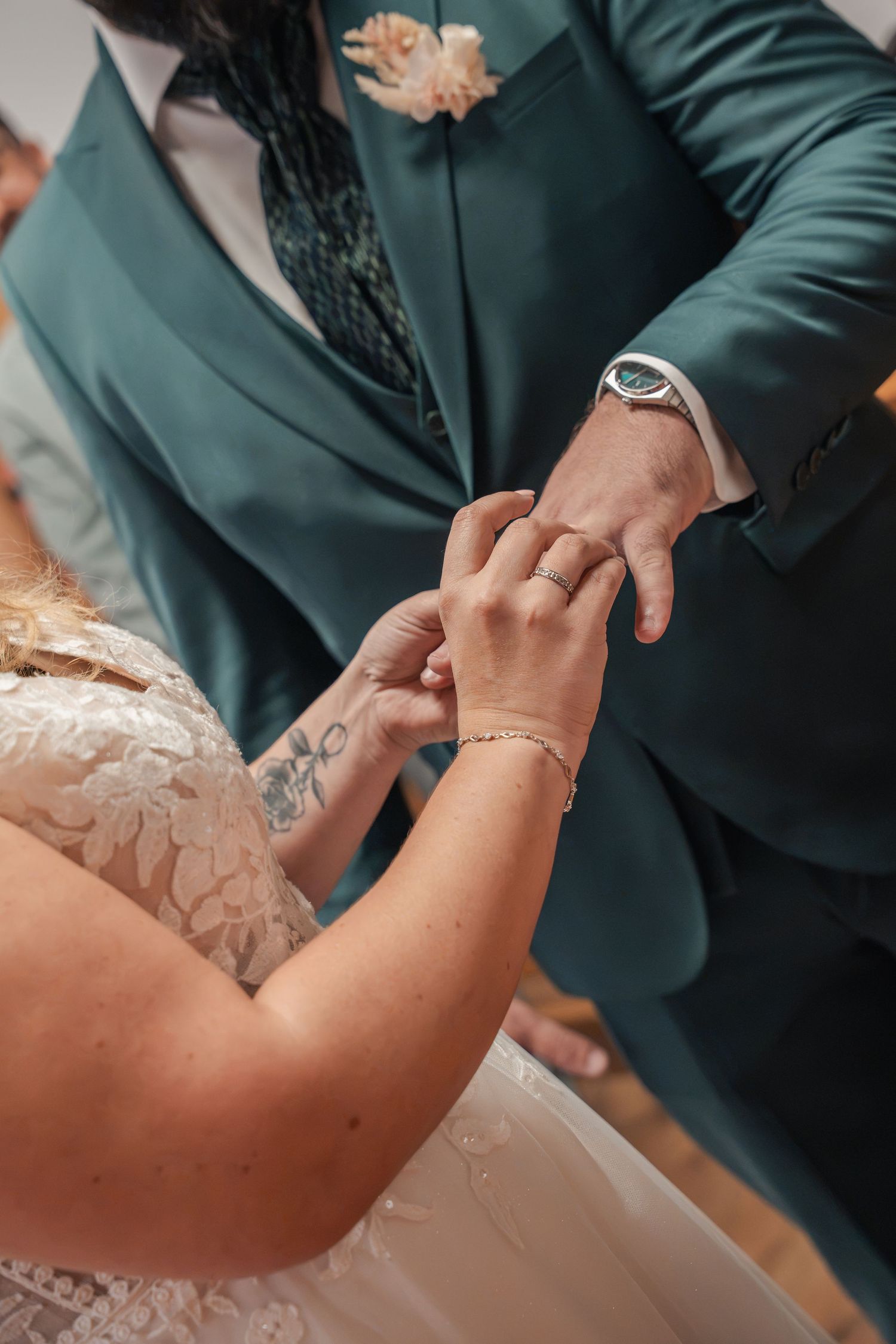 Bride in a lace wedding gown and groom in a teal suit exchange rings during a wedding ceremony, with a close view of their hands.
