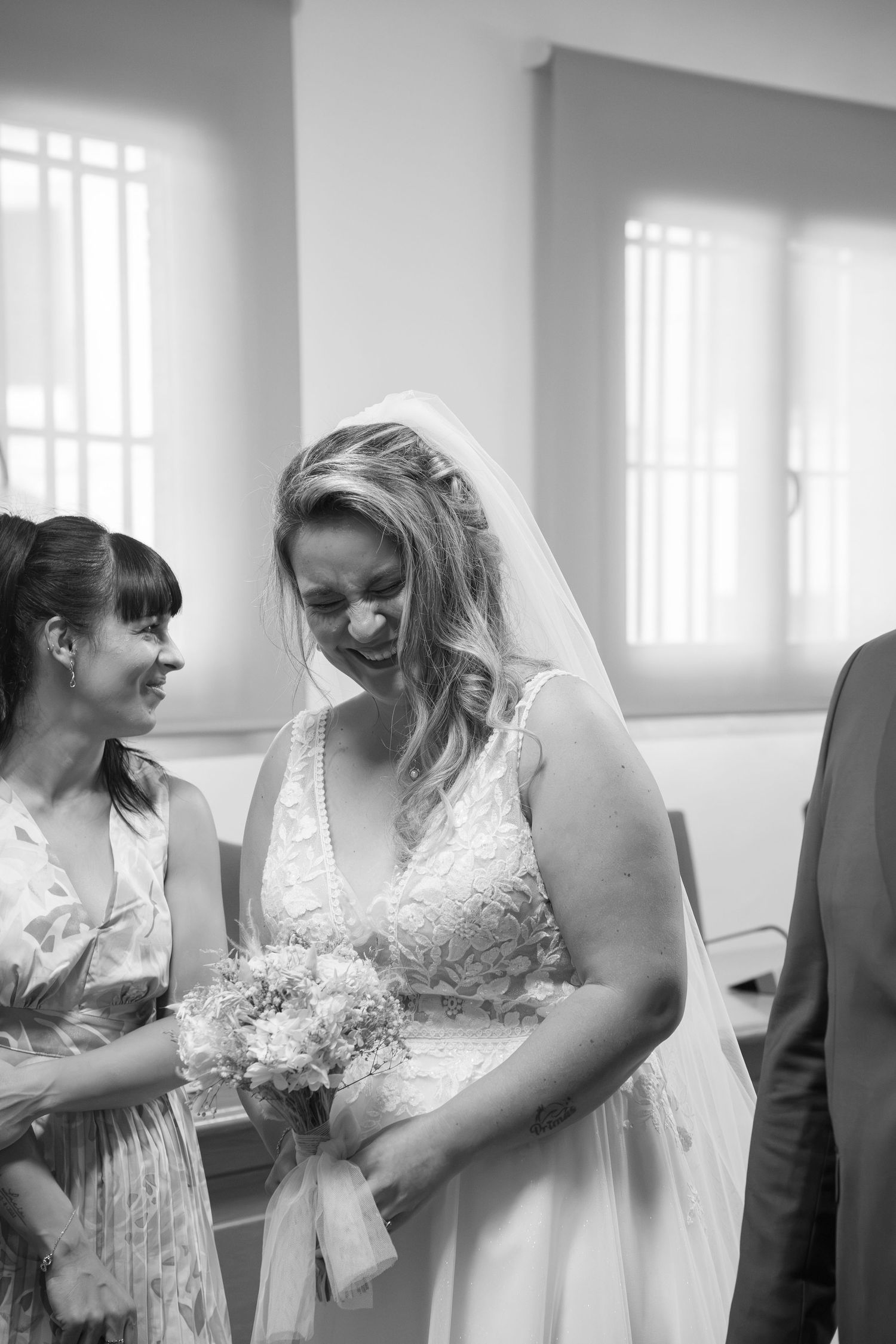 Black and white photo of a smiling bride in a lace gown and veil holding a bouquet while a woman laughs beside her at a wedding.