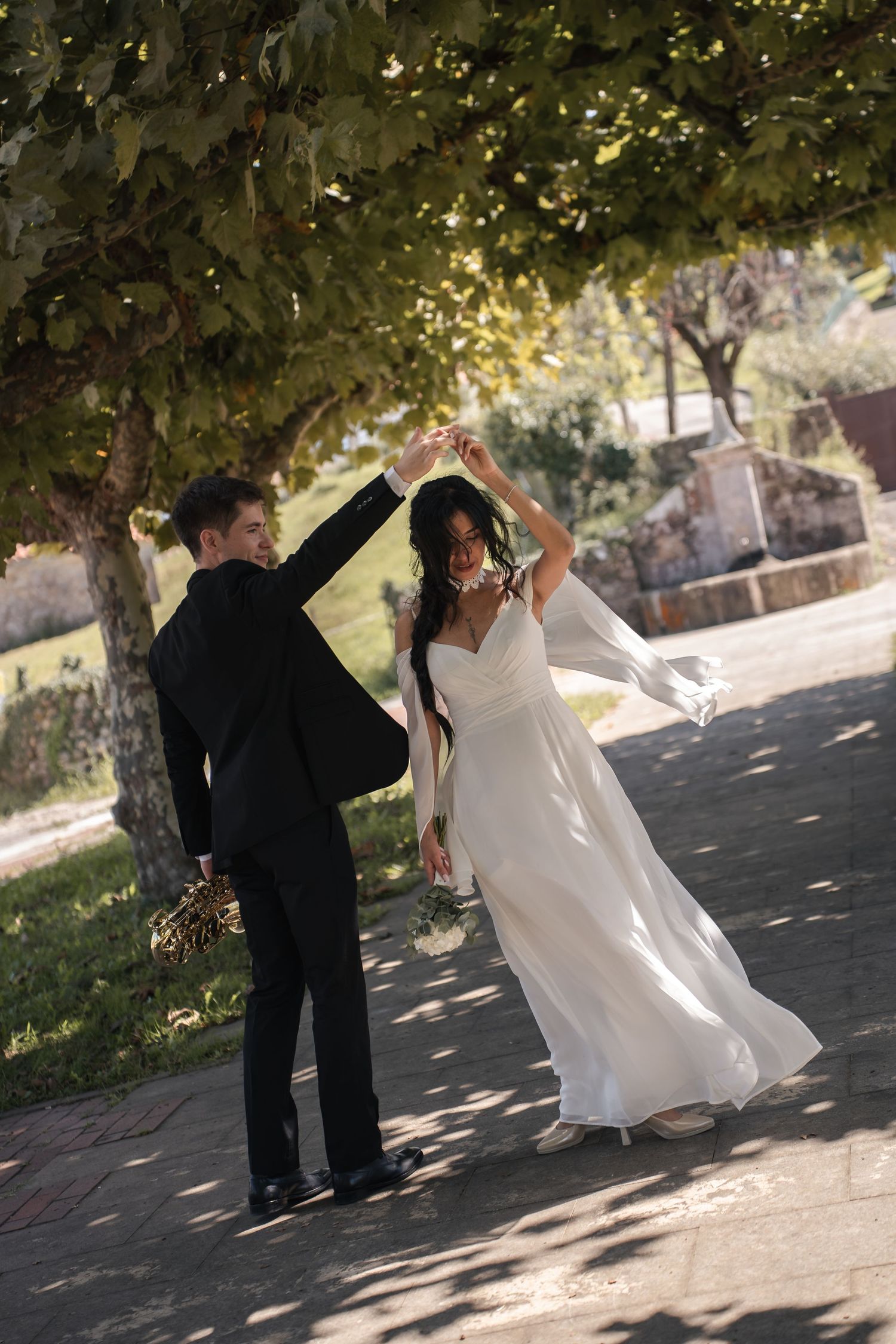 Bride in a flowing white gown and groom in a black suit dance hand in hand under leafy trees on a sunlit path.