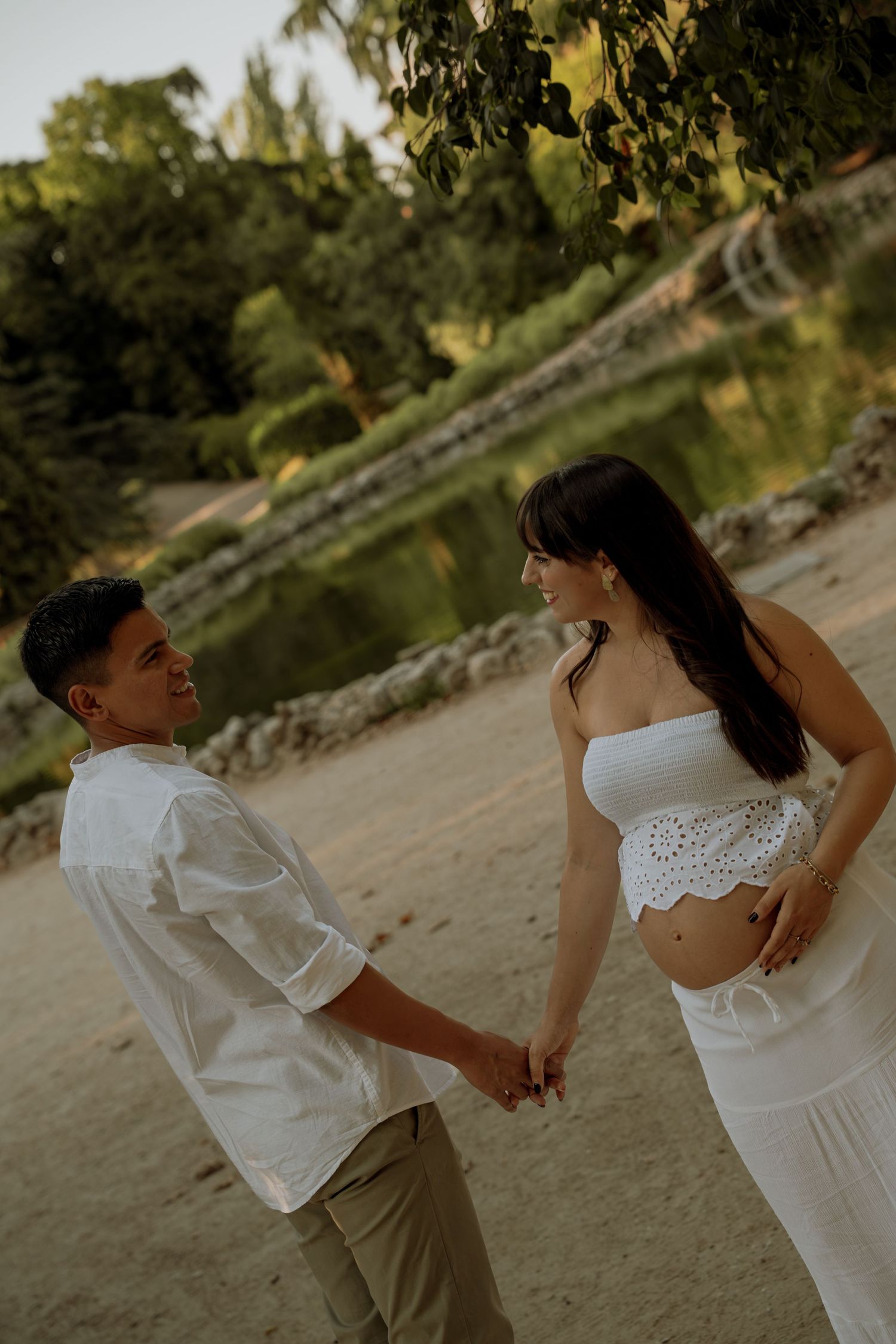 Pregnant couple holding hands near a pond, woman in white crop top and skirt, man in white shirt.