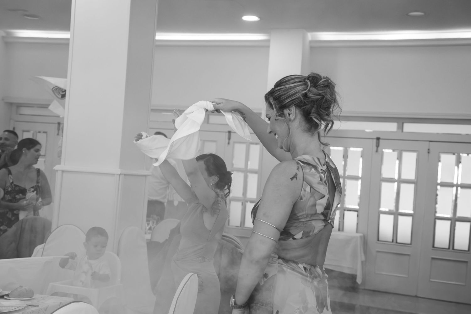 Black and white photo of a woman in an ornate dress waving a white cloth while dancing at a wedding reception with guests seated nearby.
