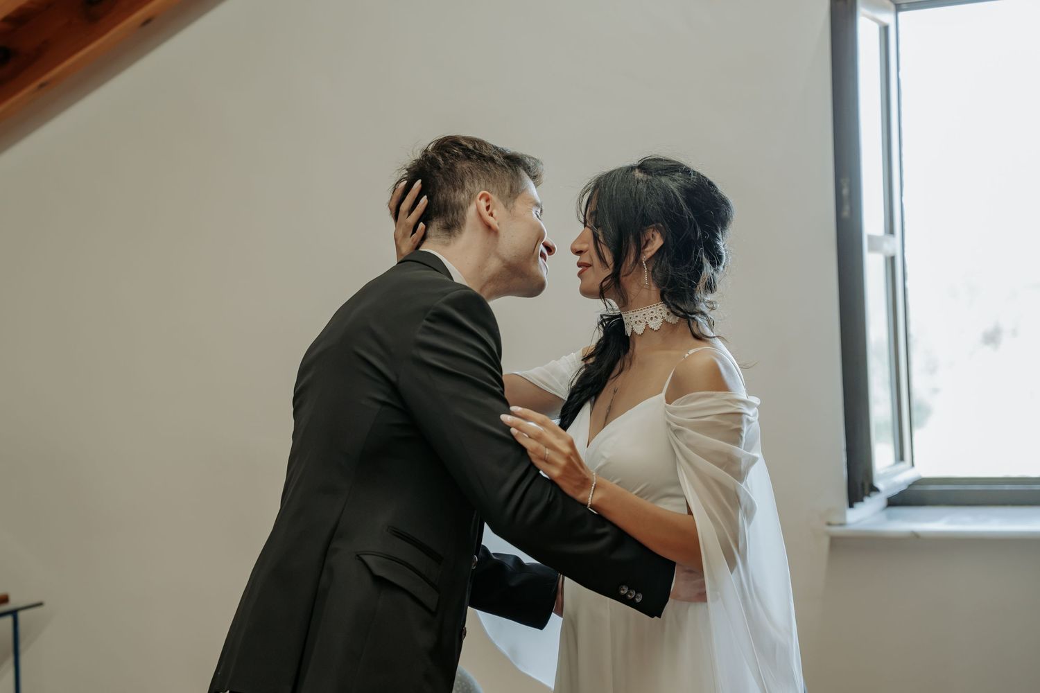 A bride in a white gown with sheer sleeves embraces a groom in a dark suit beside a bright window in an intimate wedding moment.