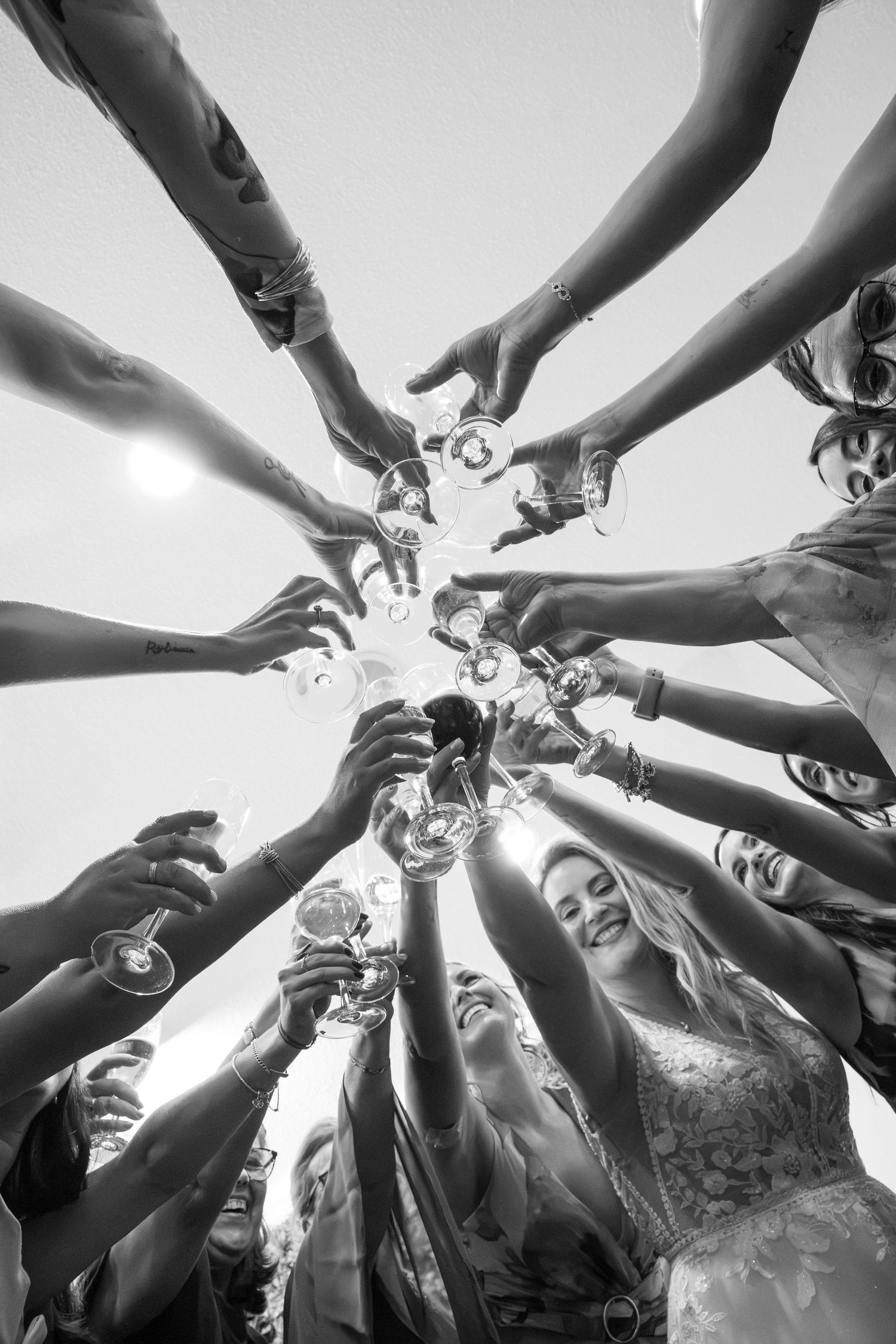 Black and white photo of wedding party toasting with champagne glasses raised together in a circle from below.