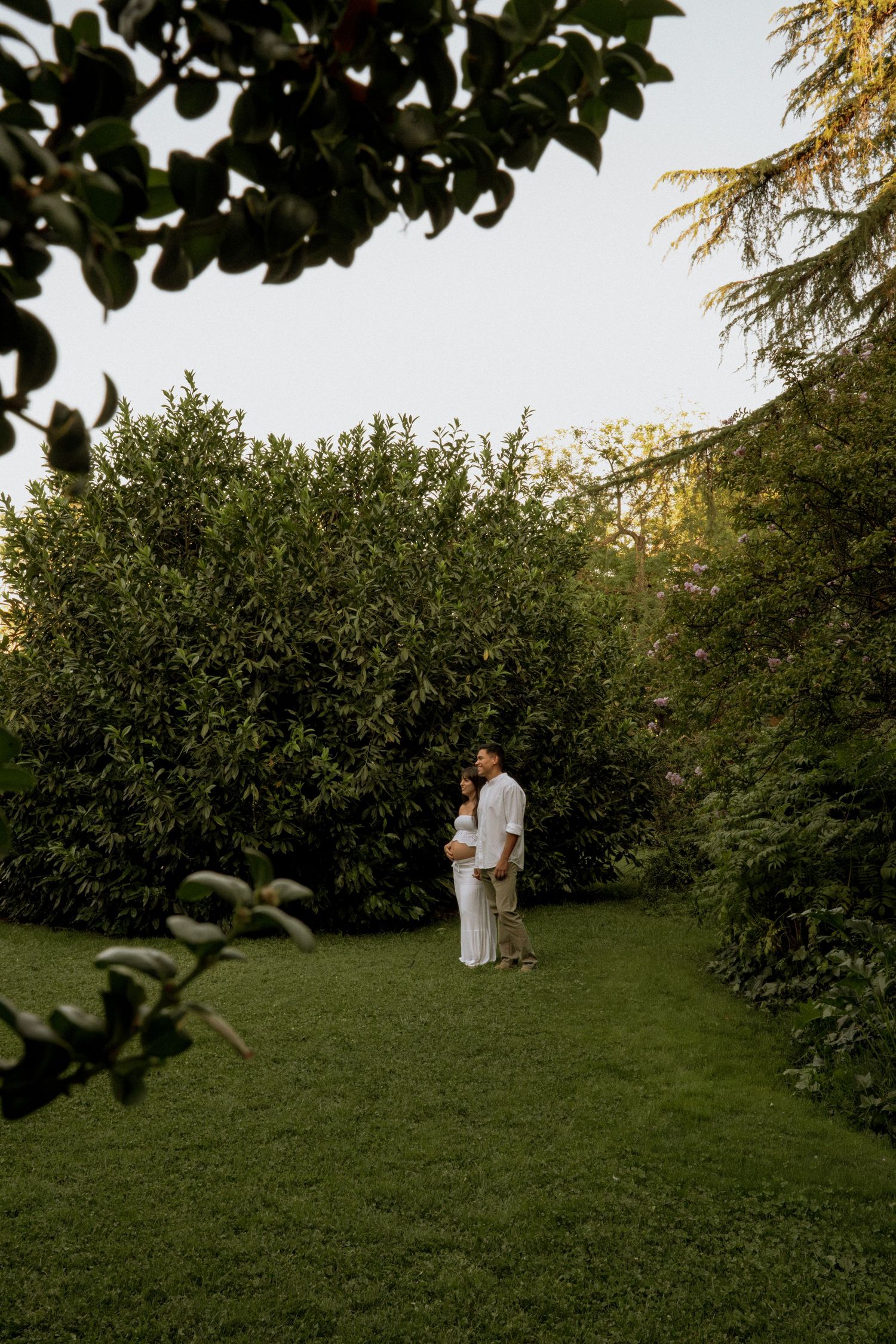 Expecting couple embracing in lush green garden corridor at golden hour, surrounded by tall trees.