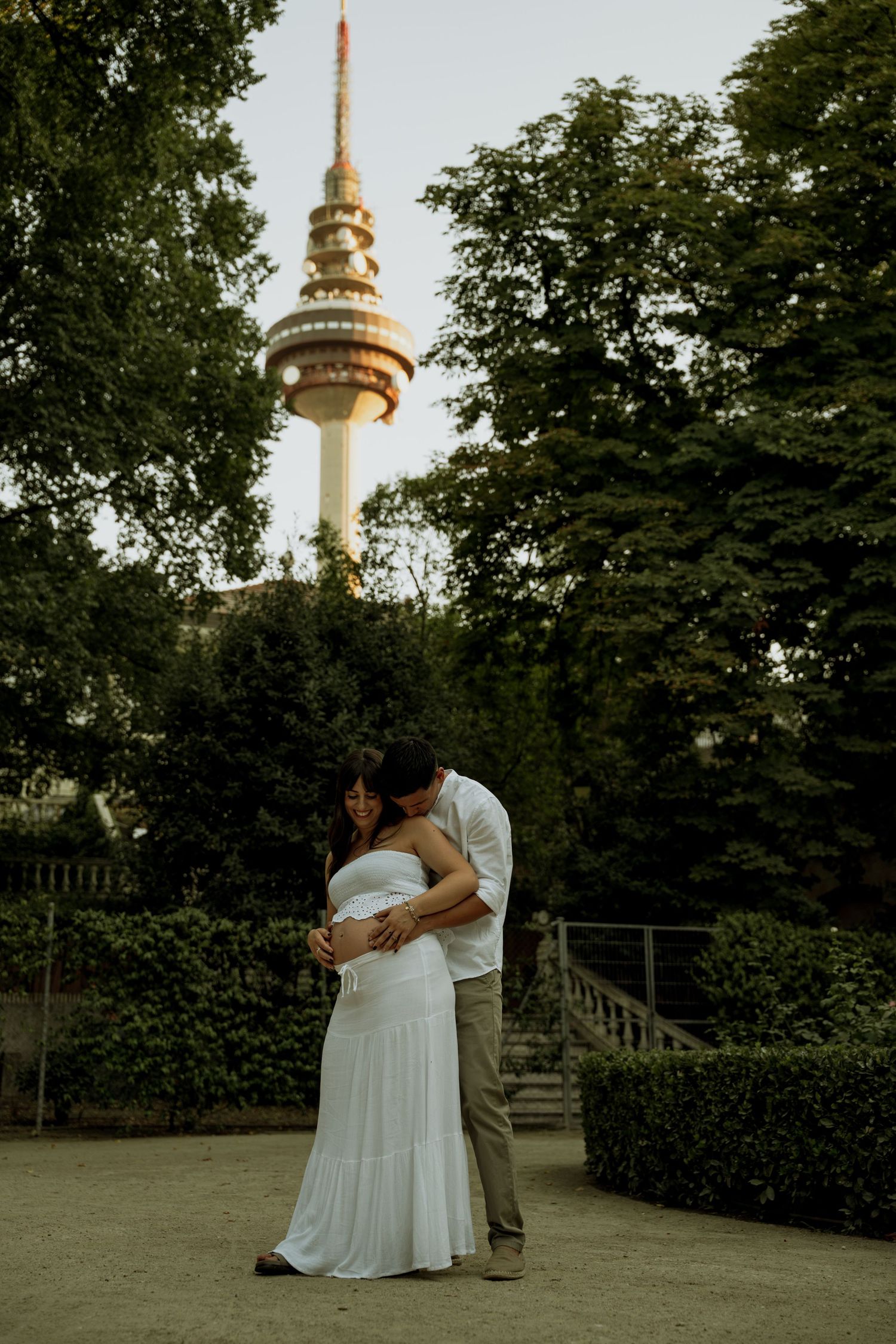 Pregnant couple embracing in park with tall communications tower visible above trees in background.
