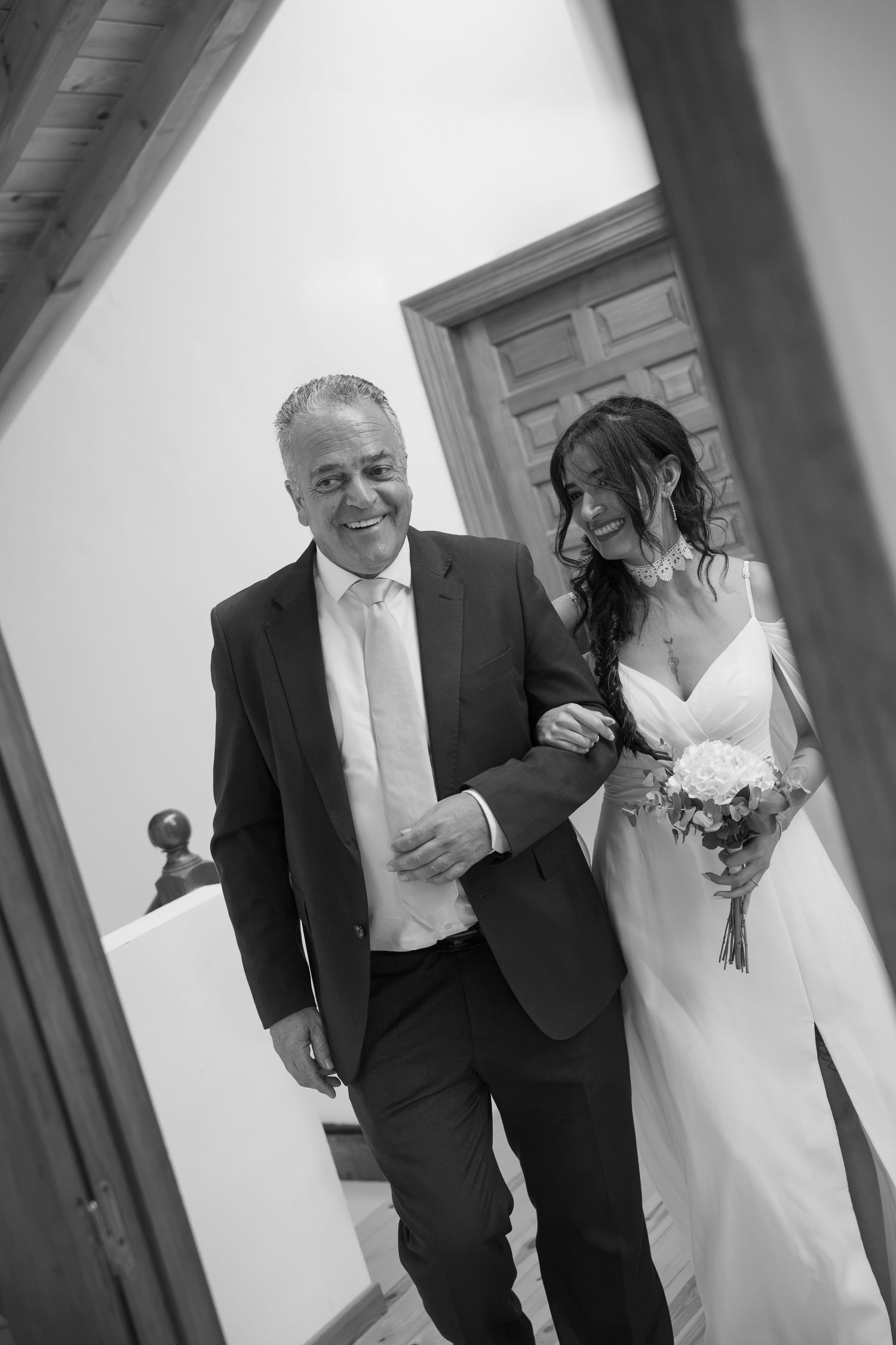 Black and white photo of a smiling bride holding bouquet walking arm in arm with a man in a suit at a wedding.