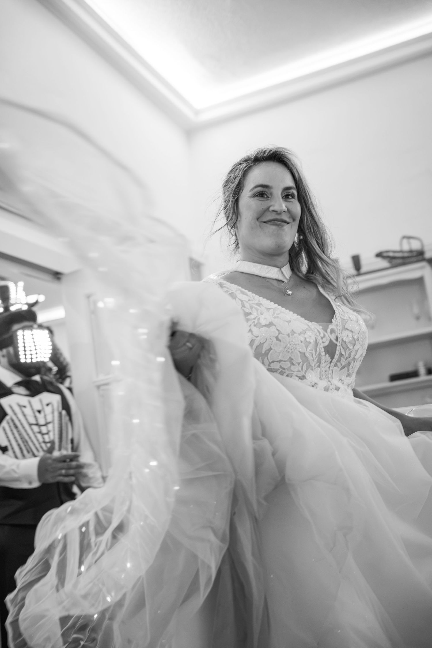 Black and white photo of a smiling bride in a lace wedding gown twirling her veil indoors.