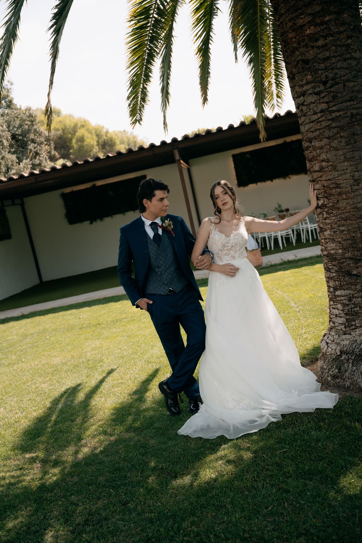 Bride and groom pose together leaning against a palm tree on green lawn at outdoor wedding venue.