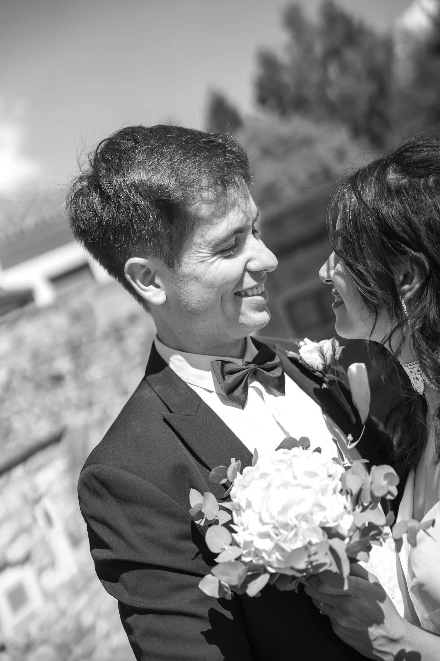 Black and white photo of a smiling groom in a bow tie gazing at his bride, who holds a white bouquet, beside a stone wall.